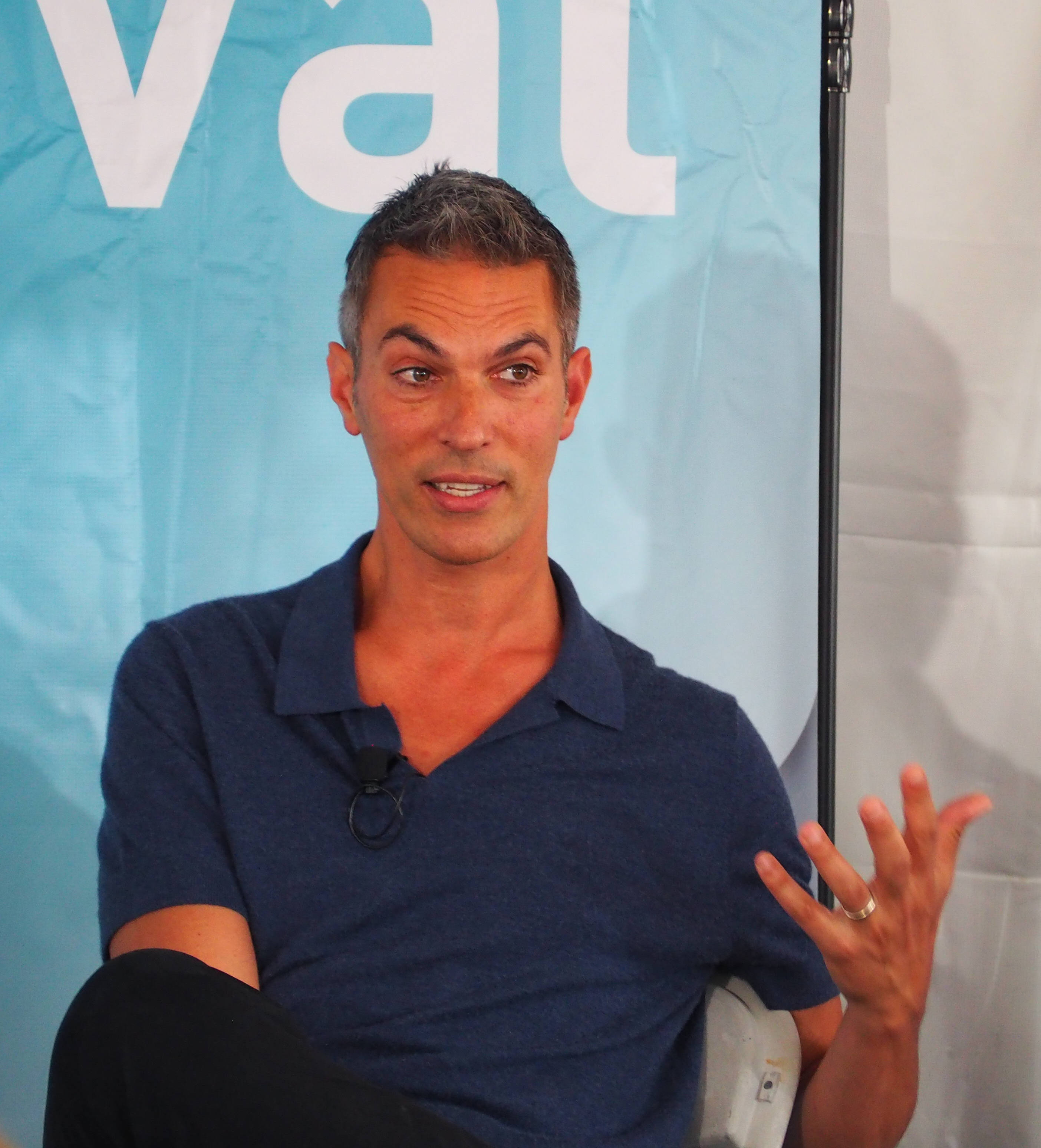 Man in navy polo shirt with lapel microphone seated and gesturing while speaking.