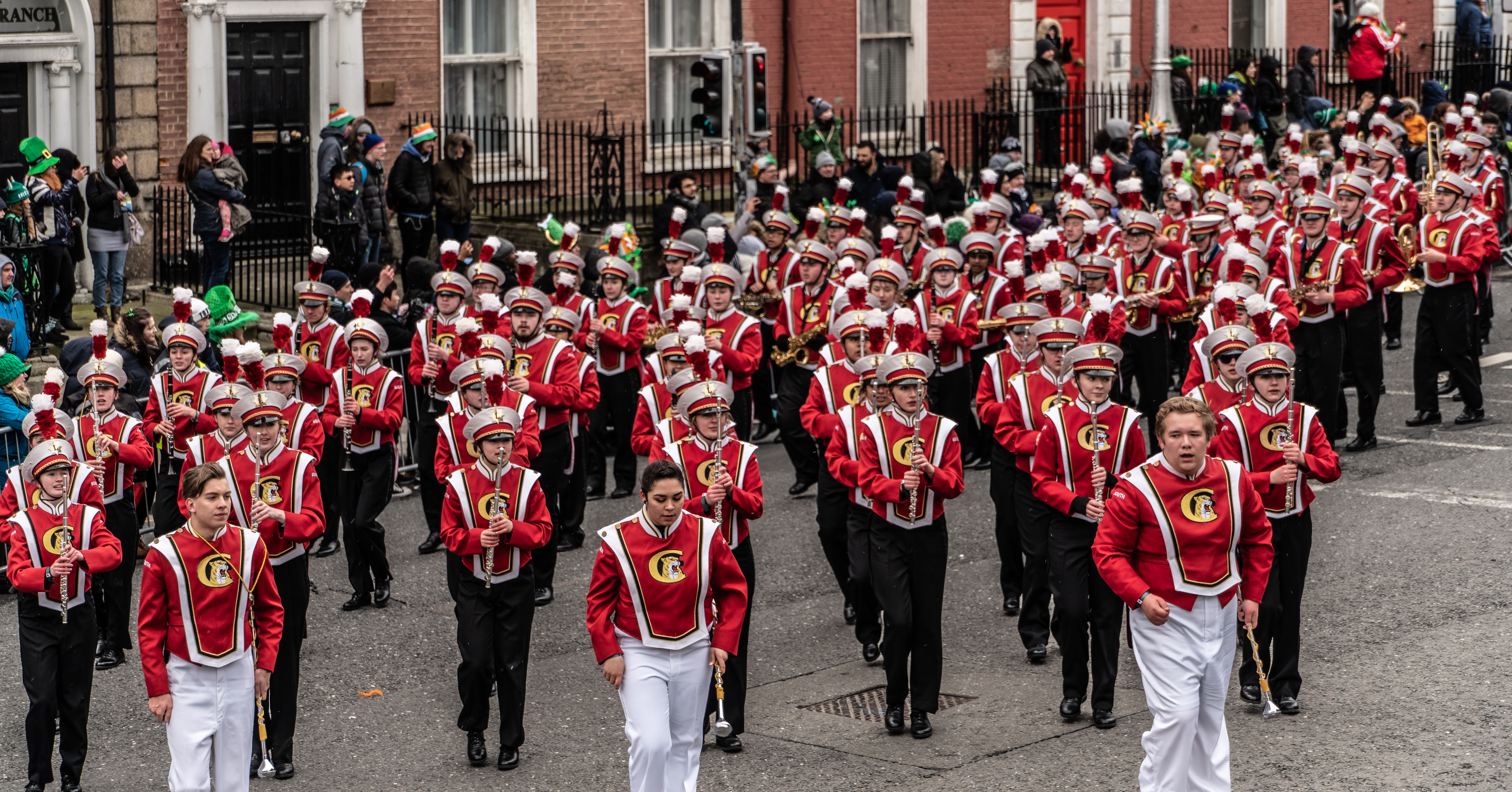 High school marching band in red and white uniforms parading down a city street.
