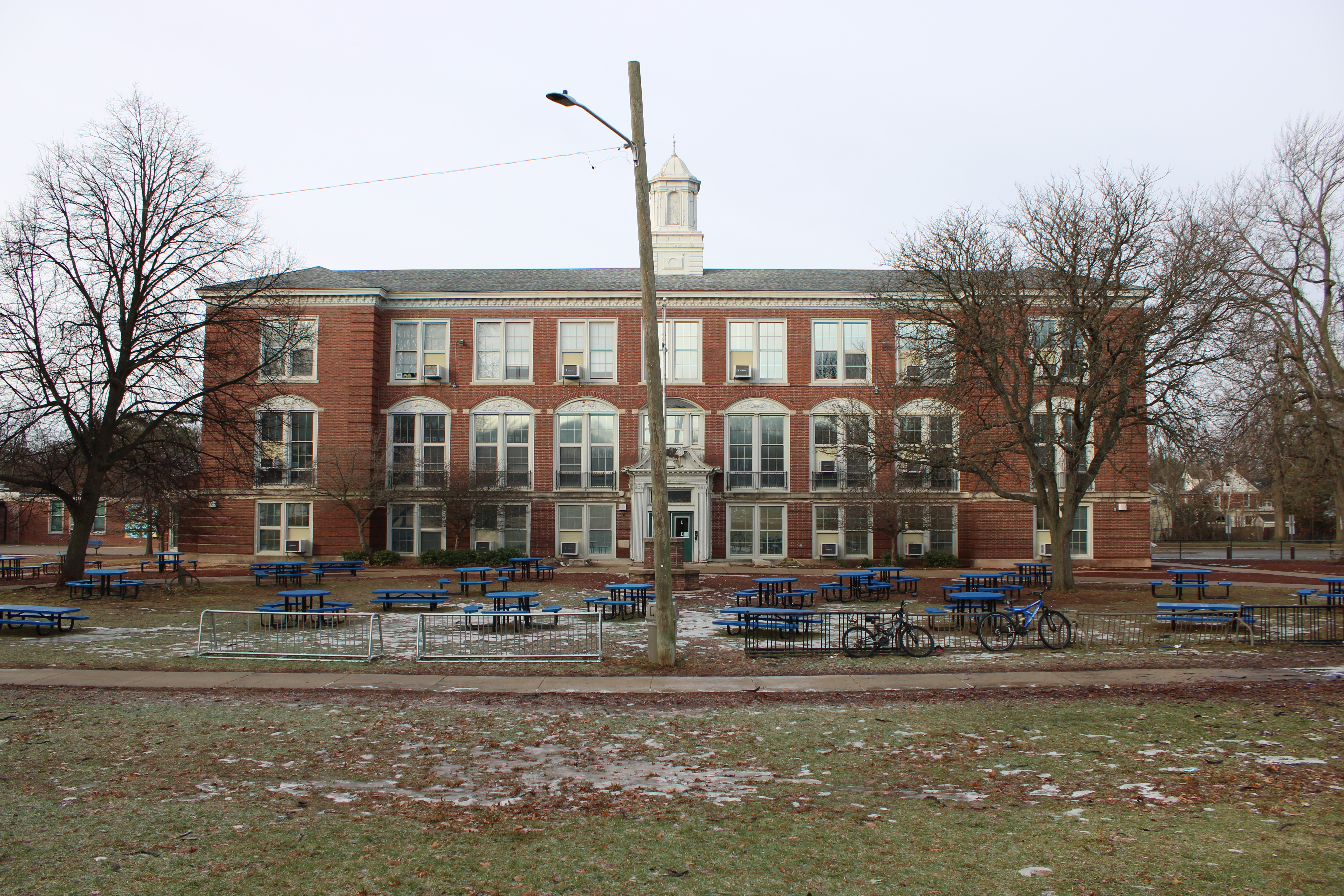 Red brick school building with white cupola, blue picnic tables, and bare winter trees.