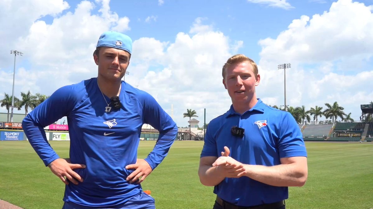 Two men in blue baseball shirts stand on a sunny field with palm trees behind them.