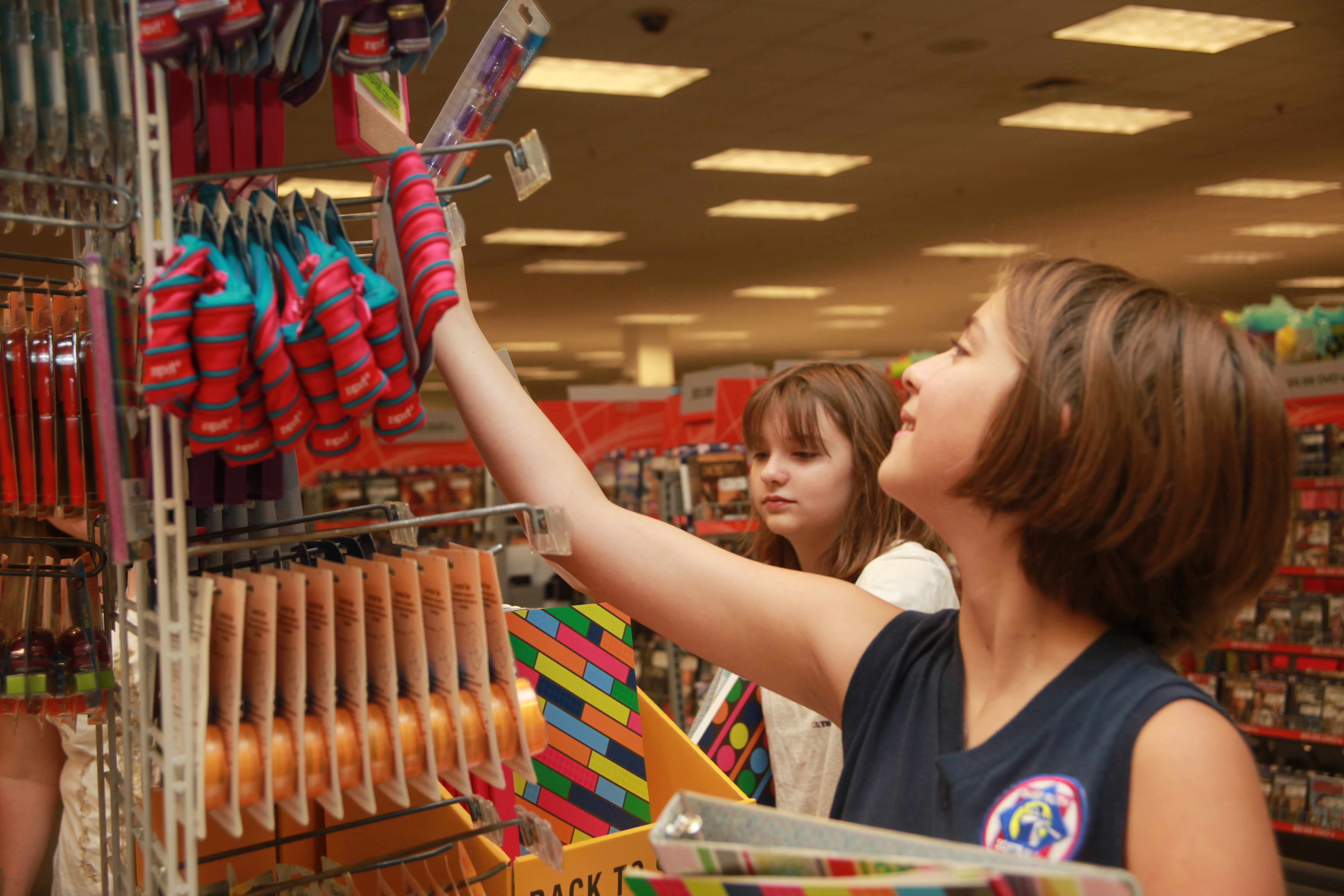Two girls reach for items on a brightly lit retail store display rack near a shopping cart.