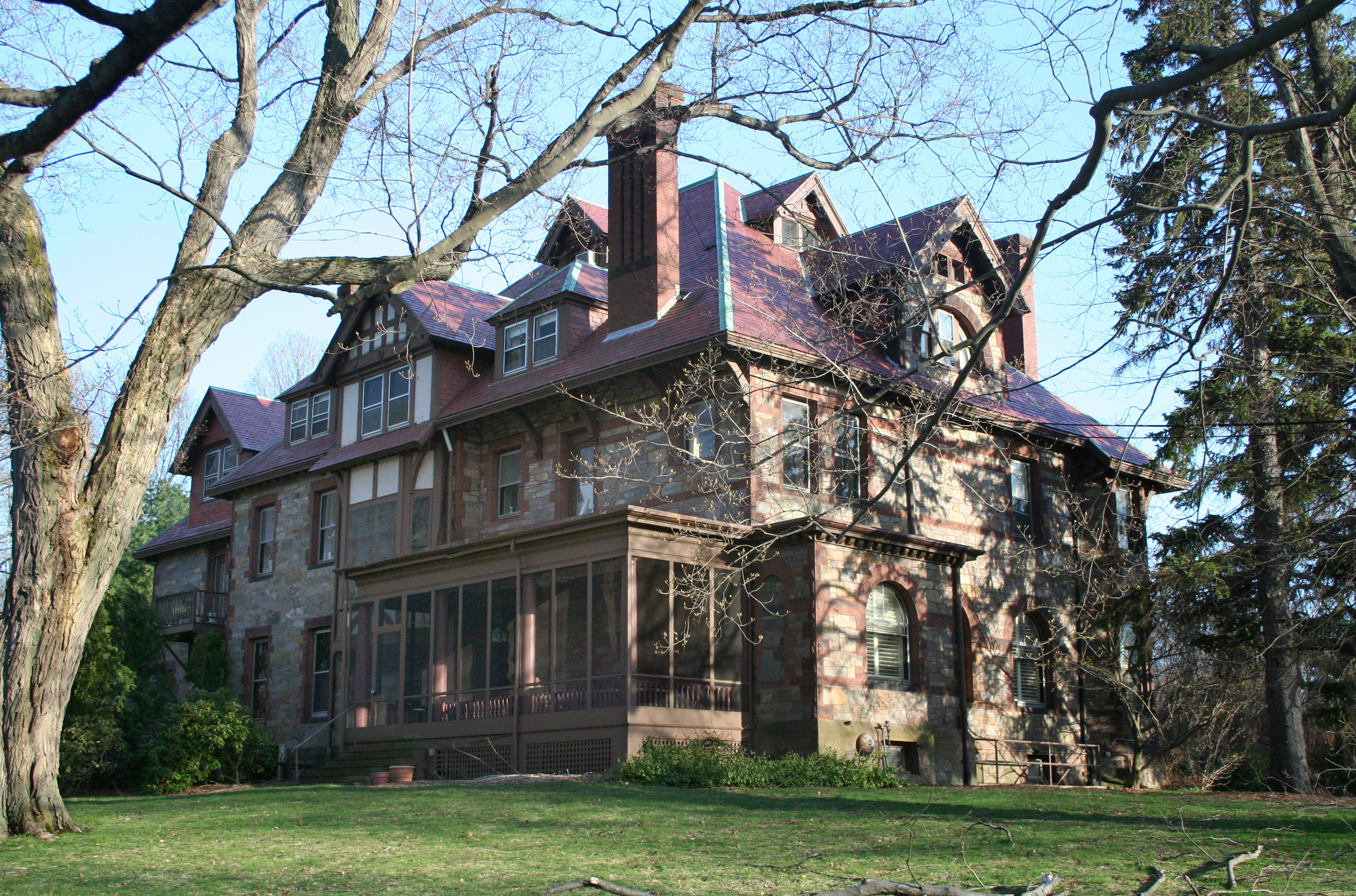 A large Victorian stone mansion with a red roof and wraparound porch surrounded by trees.