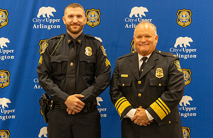 Two uniformed police officers stand side by side in front of a blue city-branded backdrop.