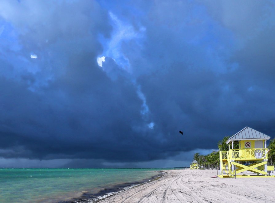 Dark storm clouds loom over a white sand beach with turquoise water and a yellow lifeguard stand.