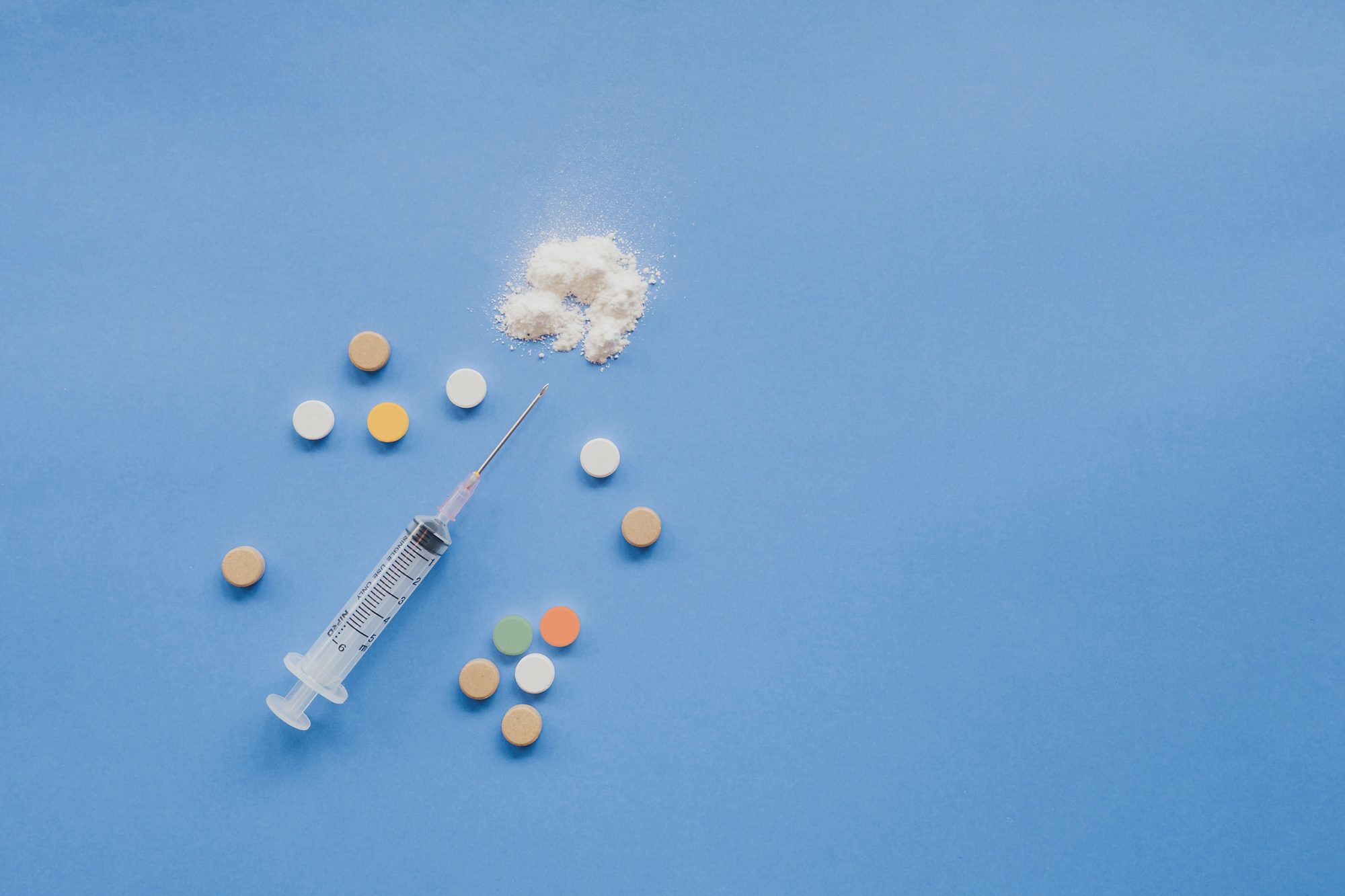 From above of syringe with multicolored pills and powder placed on blue background