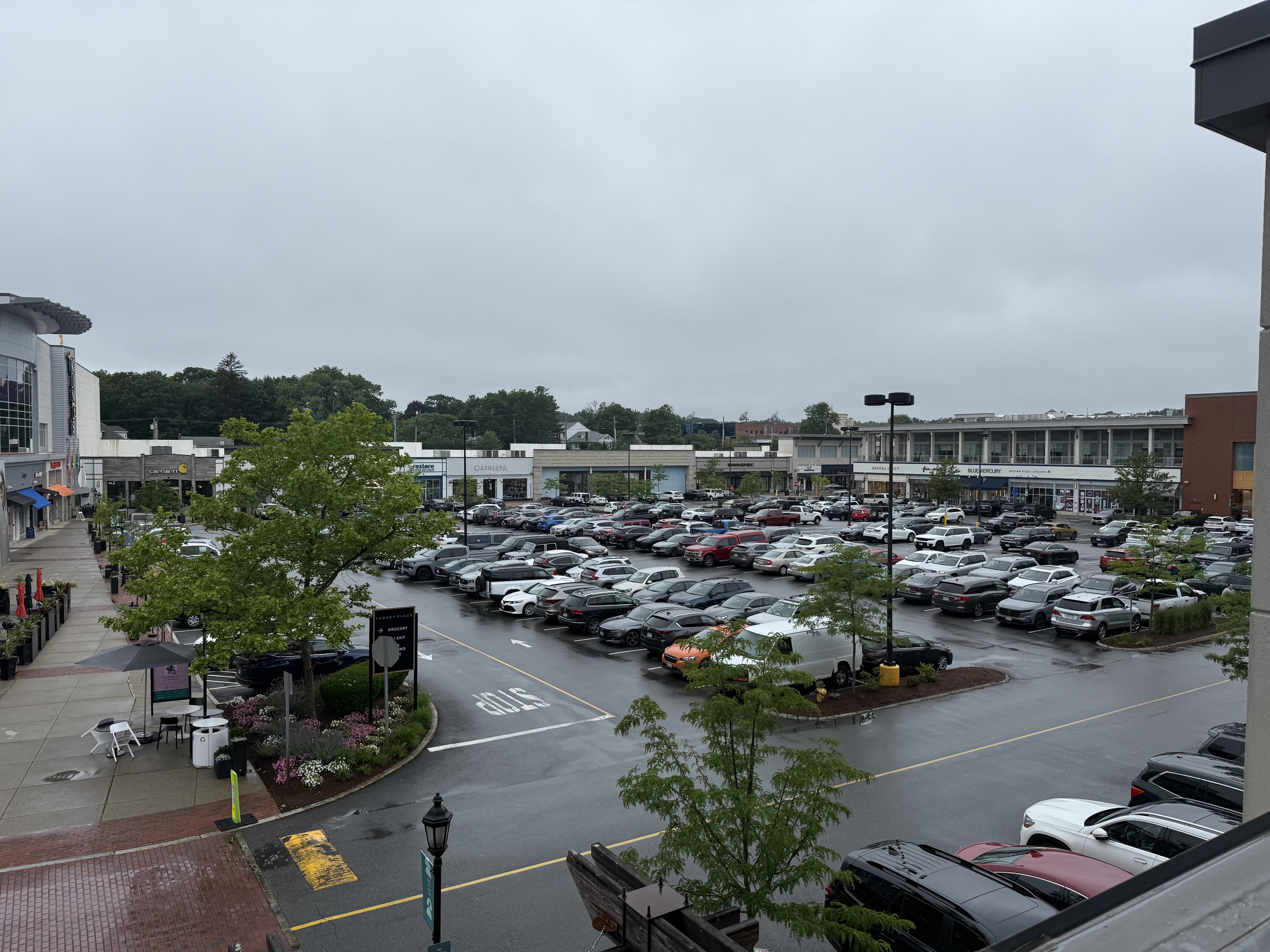 Elevated view of a large open-air shopping center parking lot with retail storefronts and trees.