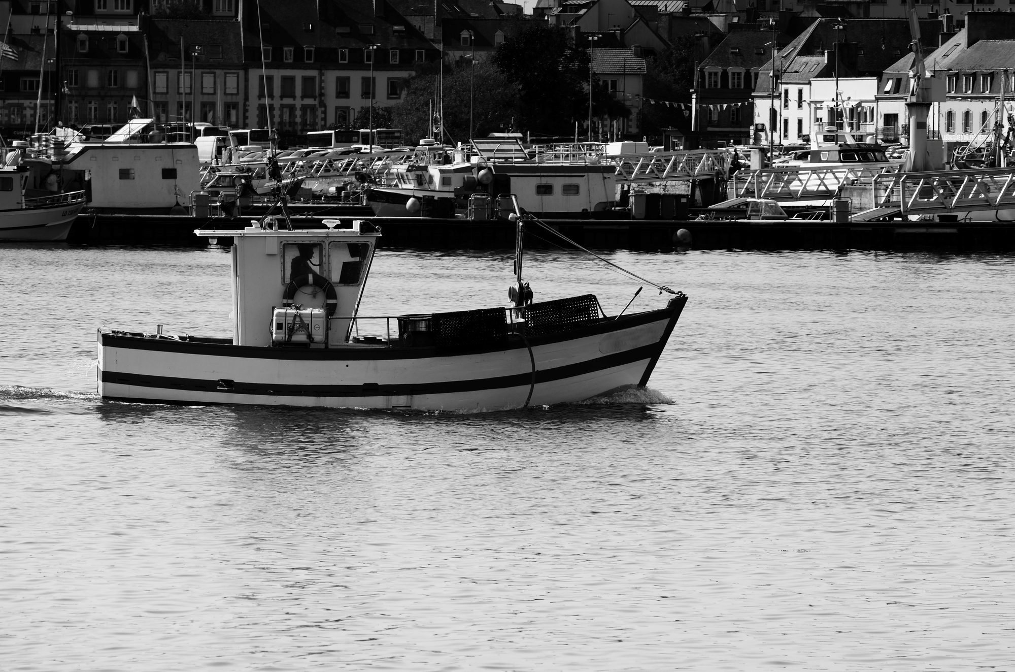 A small fishing boat in black and white sailing on a calm sea against a backdrop of a coastal town.