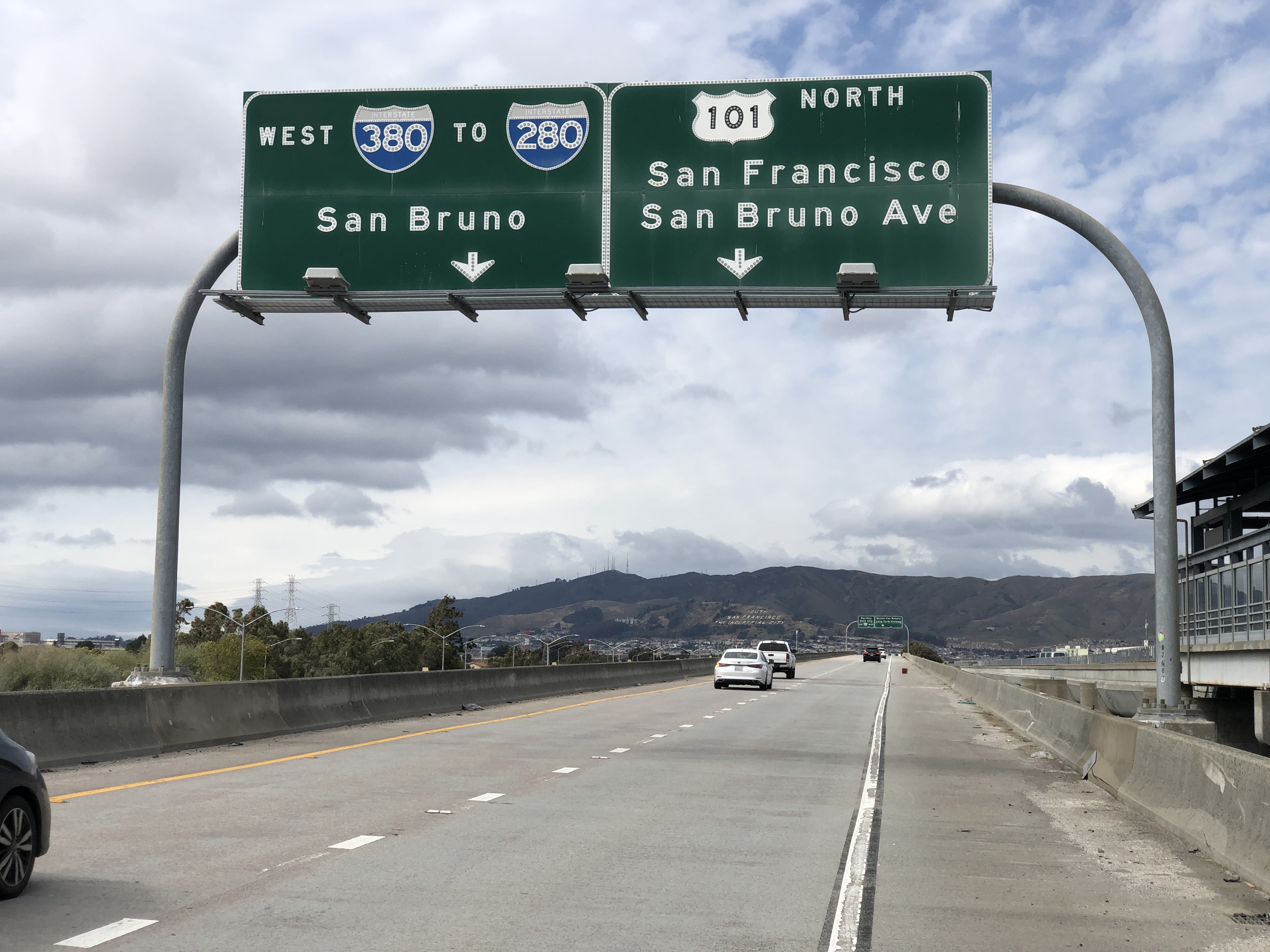 Overhead highway signs on northbound US-101 with lanes and hills in background.