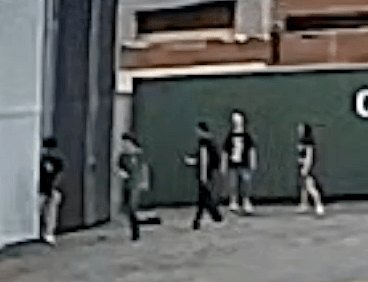 Surveillance camera image showing several people walking near a large green building.