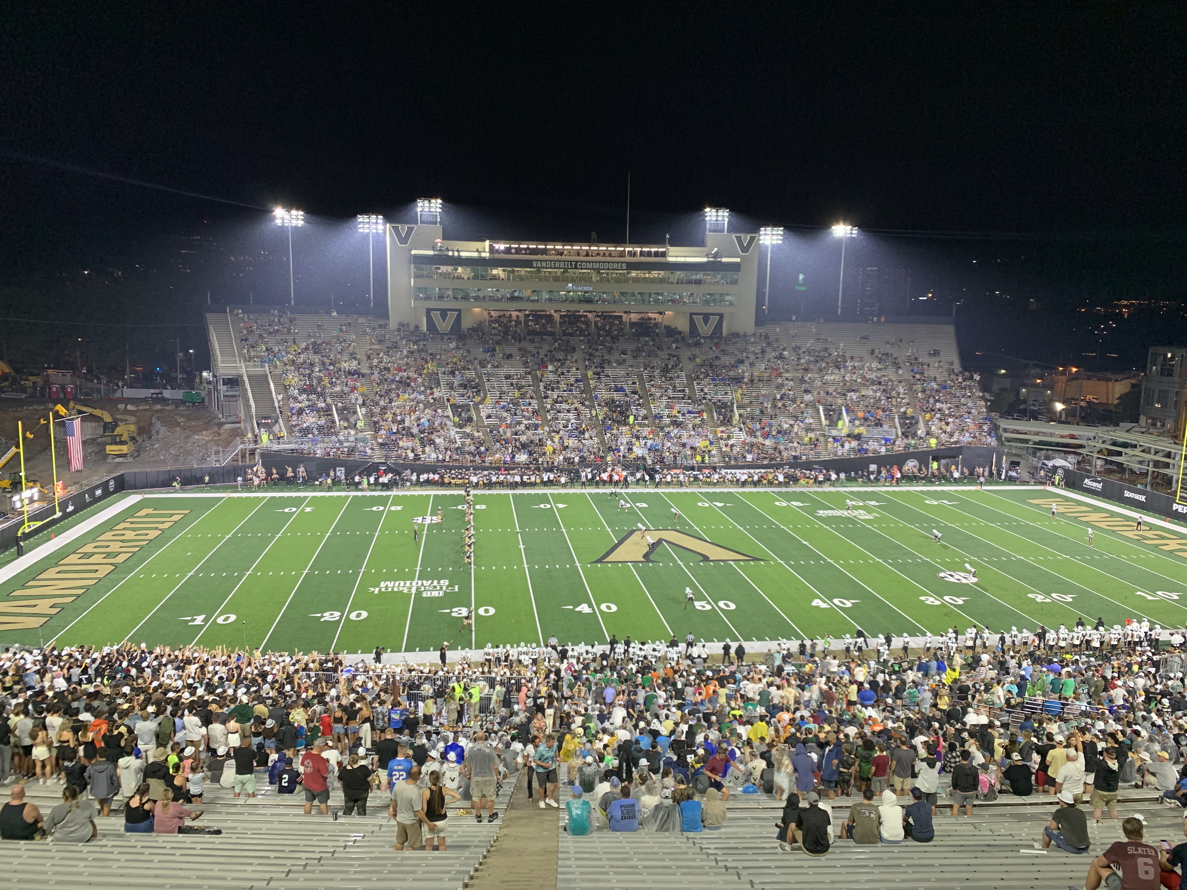 Packed college football stadium at night with bright green field and Vanderbilt branding visible.