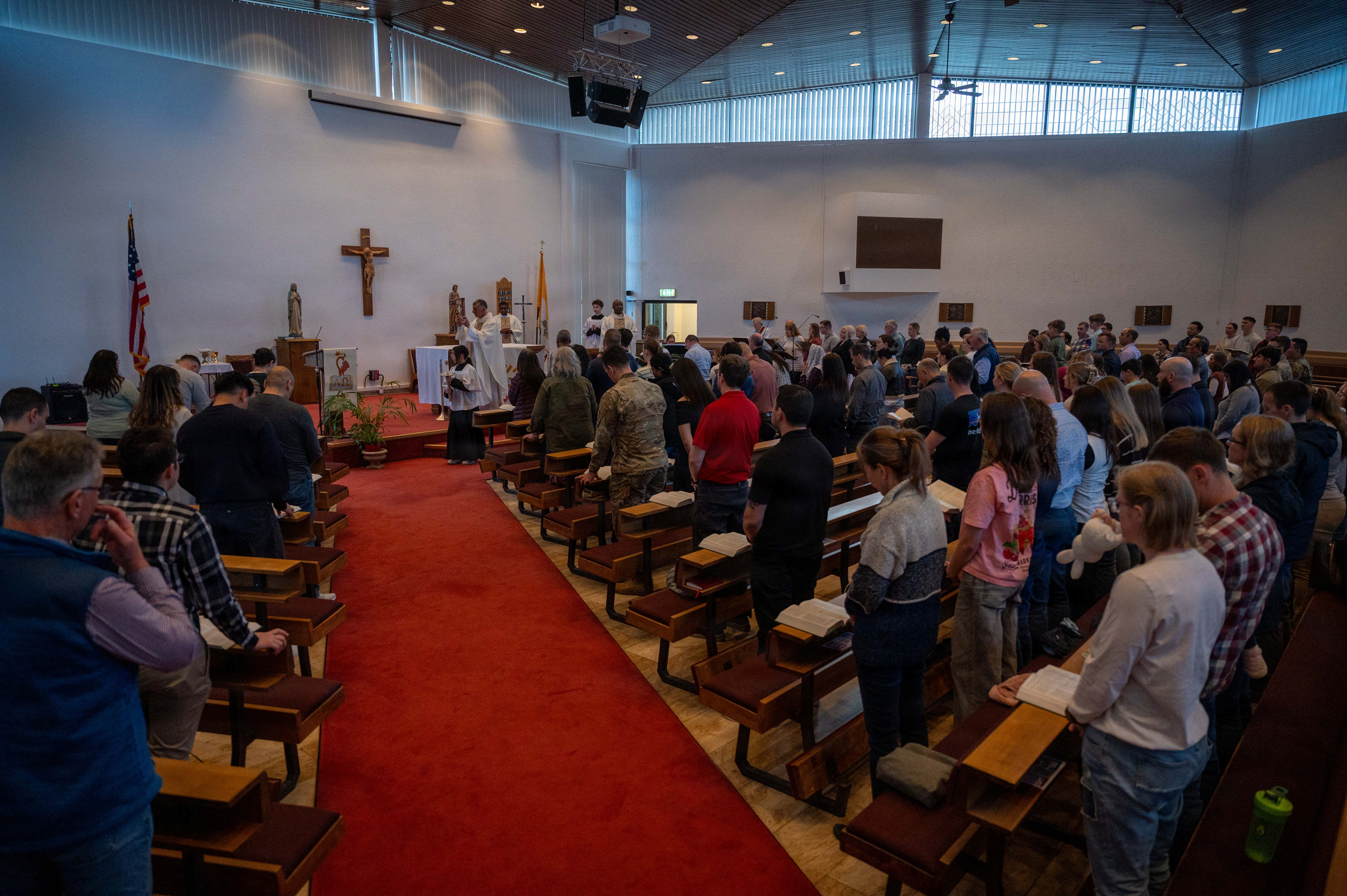Congregation standing in prayer inside a modern chapel with a crucifix at the front.