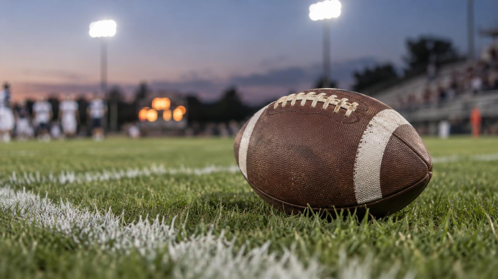 An American football resting on green grass near a white yard line under stadium lights.