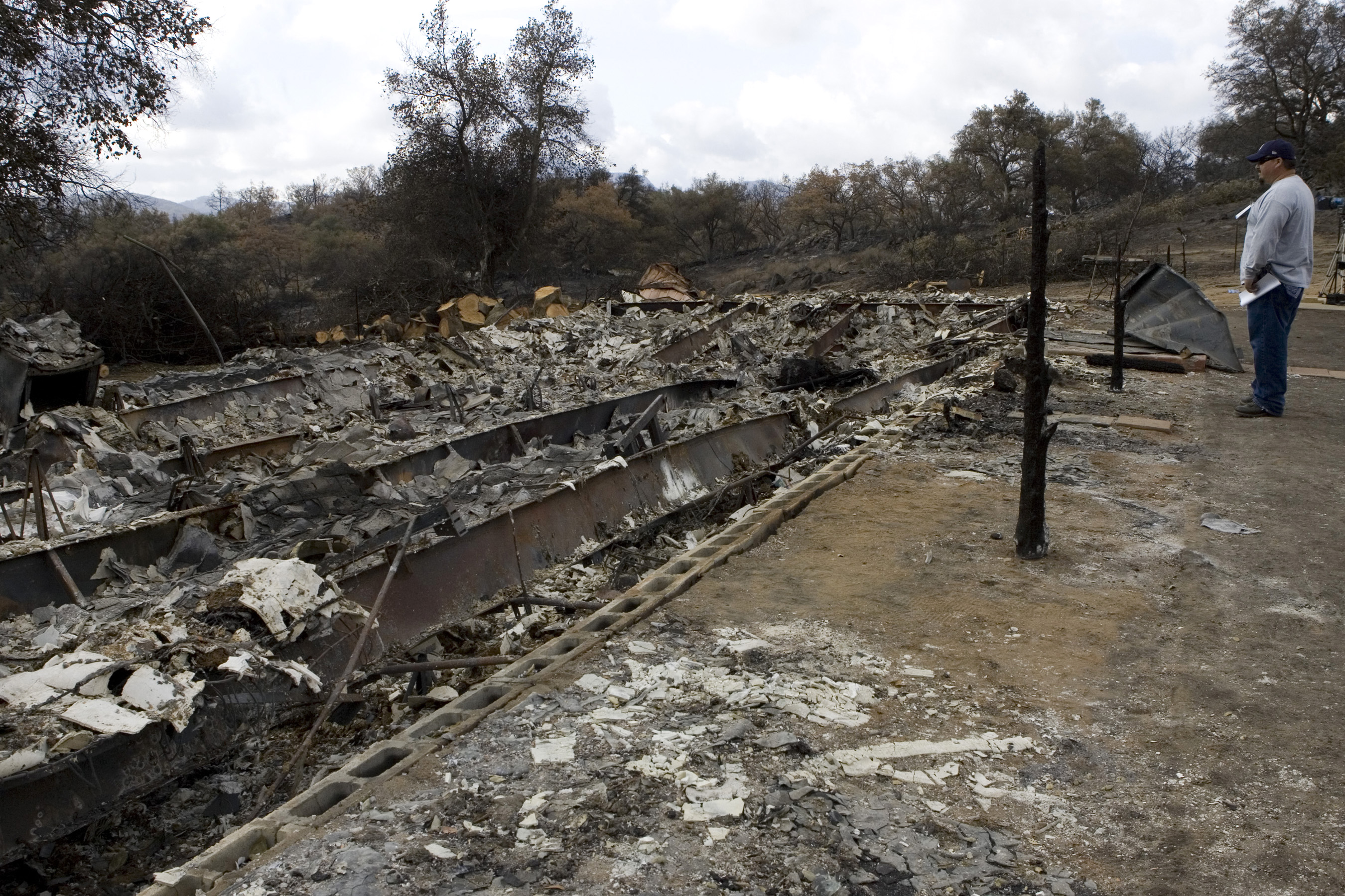 A person stands beside the charred ruins of a home destroyed by wildfire.