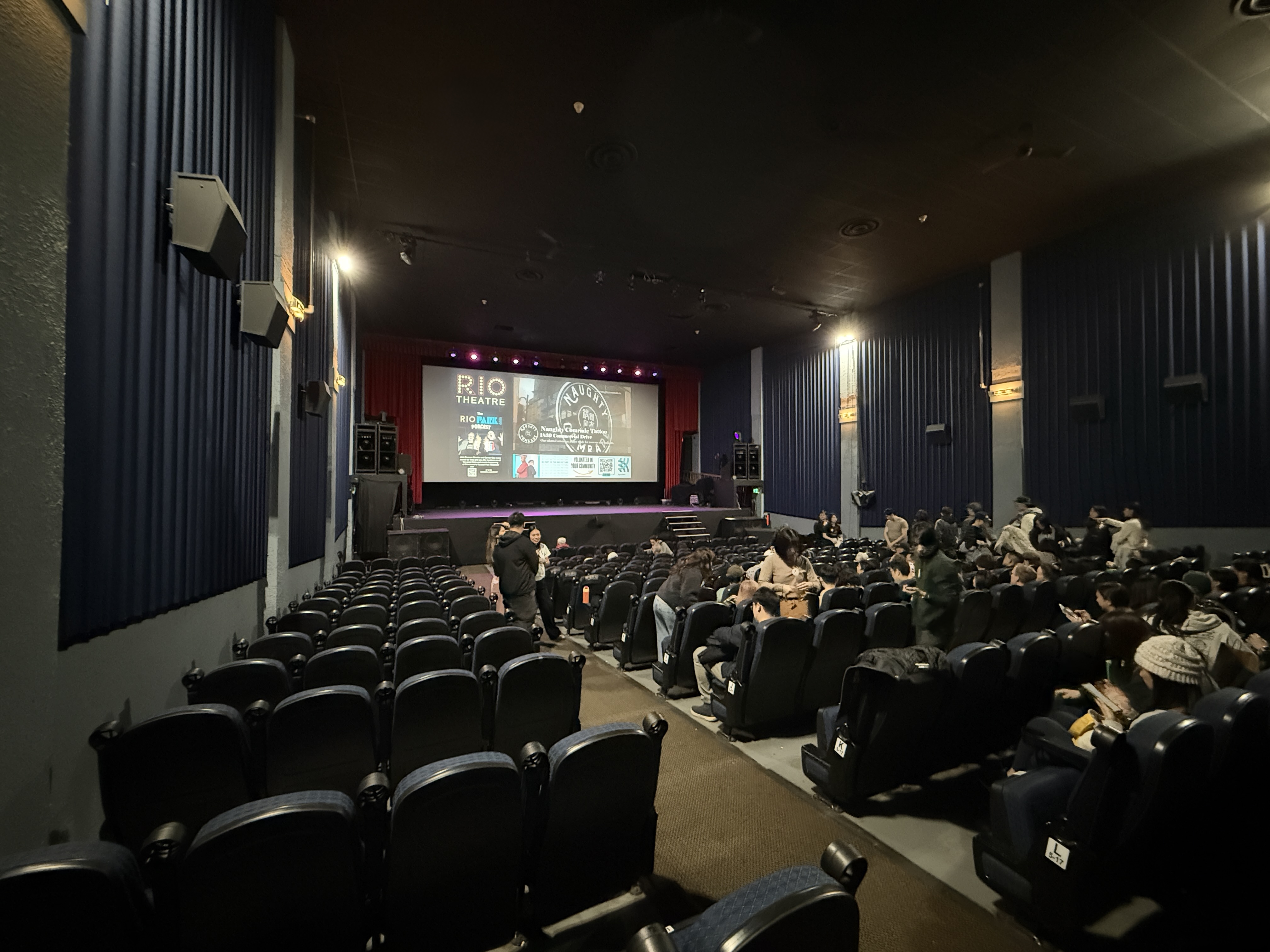 Theatre auditorium interior with rows of dark seats and a lit stage screen.