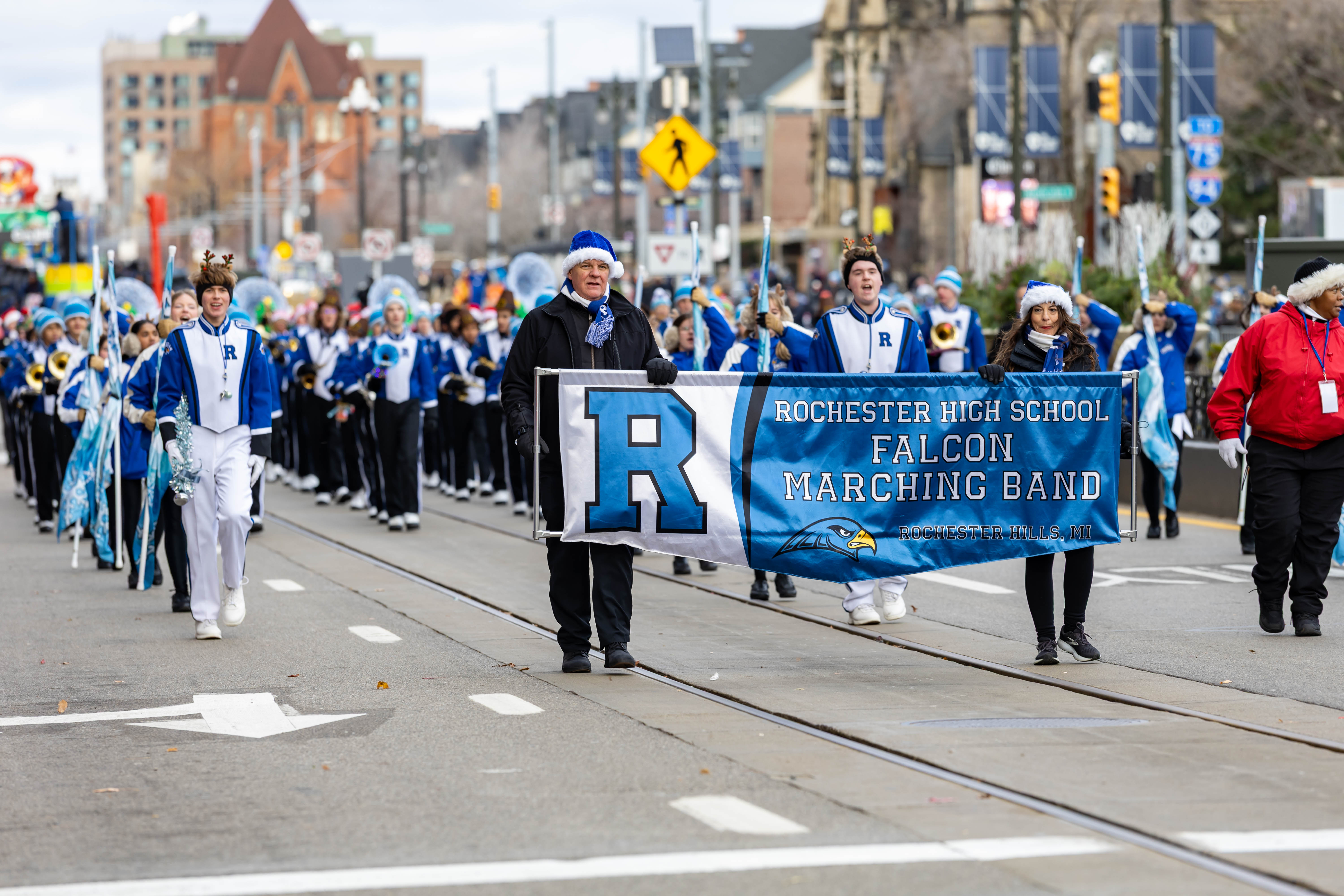Marching band members in blue and white uniforms carrying a banner on a city street.
