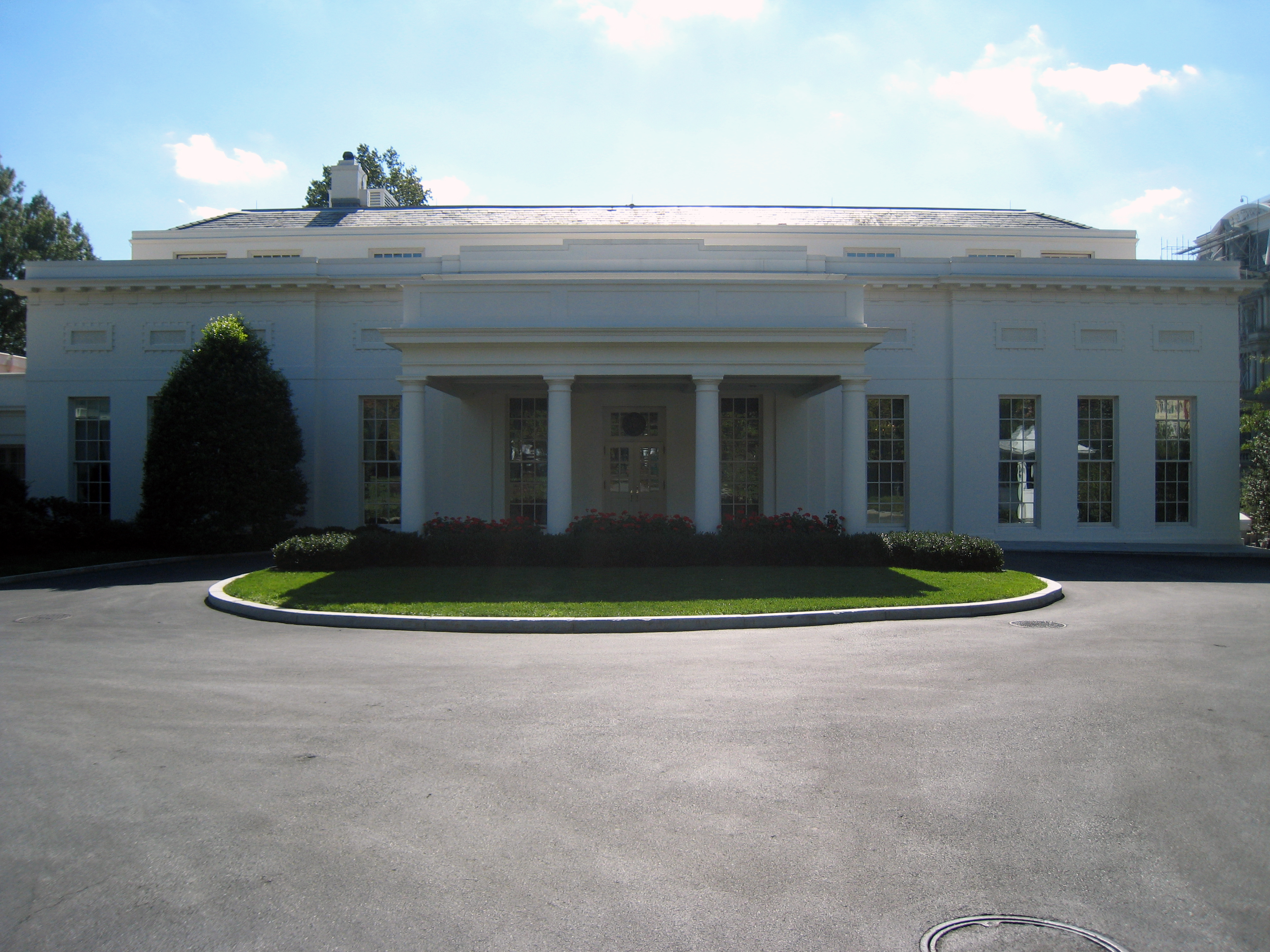 Daytime exterior of the White House West Wing with white facade and green hedges.