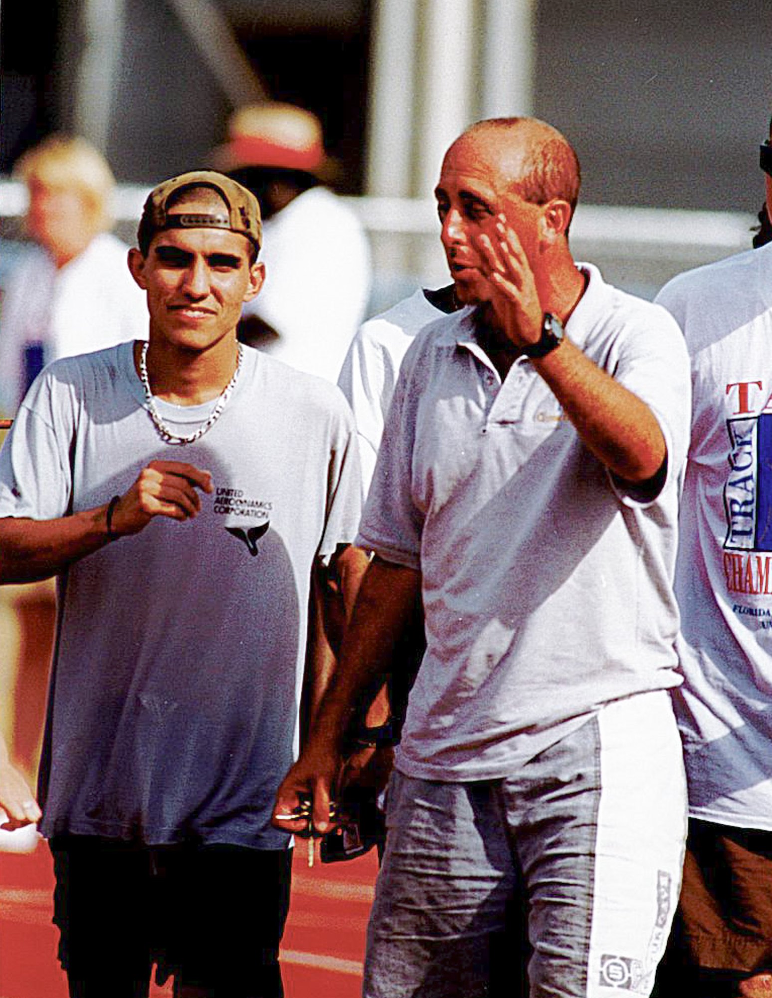 Two men walk together at a track and field facility, one gesturing.