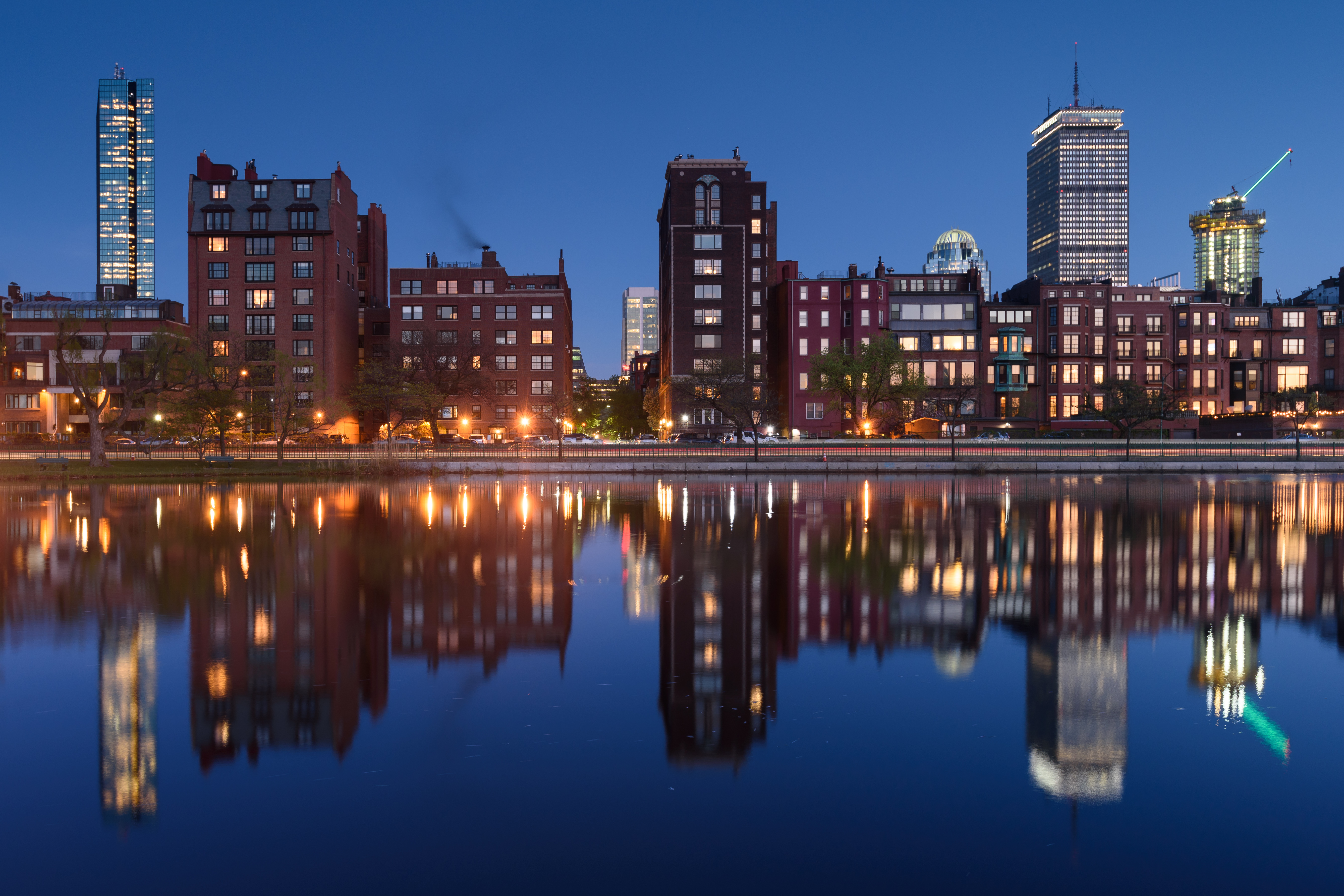 City skyline and brick buildings reflected in calm river water at dusk.