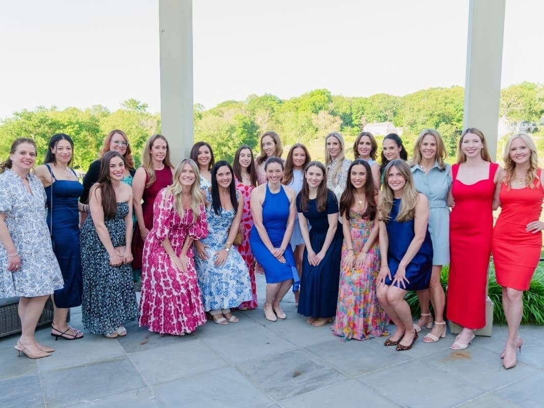 A large group of women in colorful dresses posing together on a covered porch at an outdoor event.