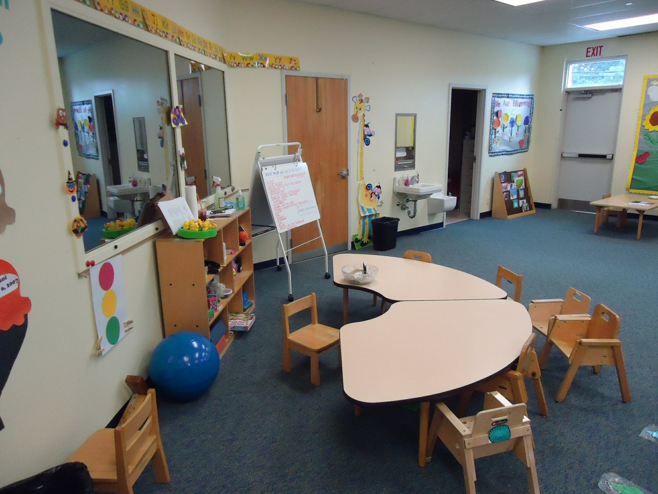 Empty preschool classroom with small chairs, a table, whiteboard, and colorful wall decorations.