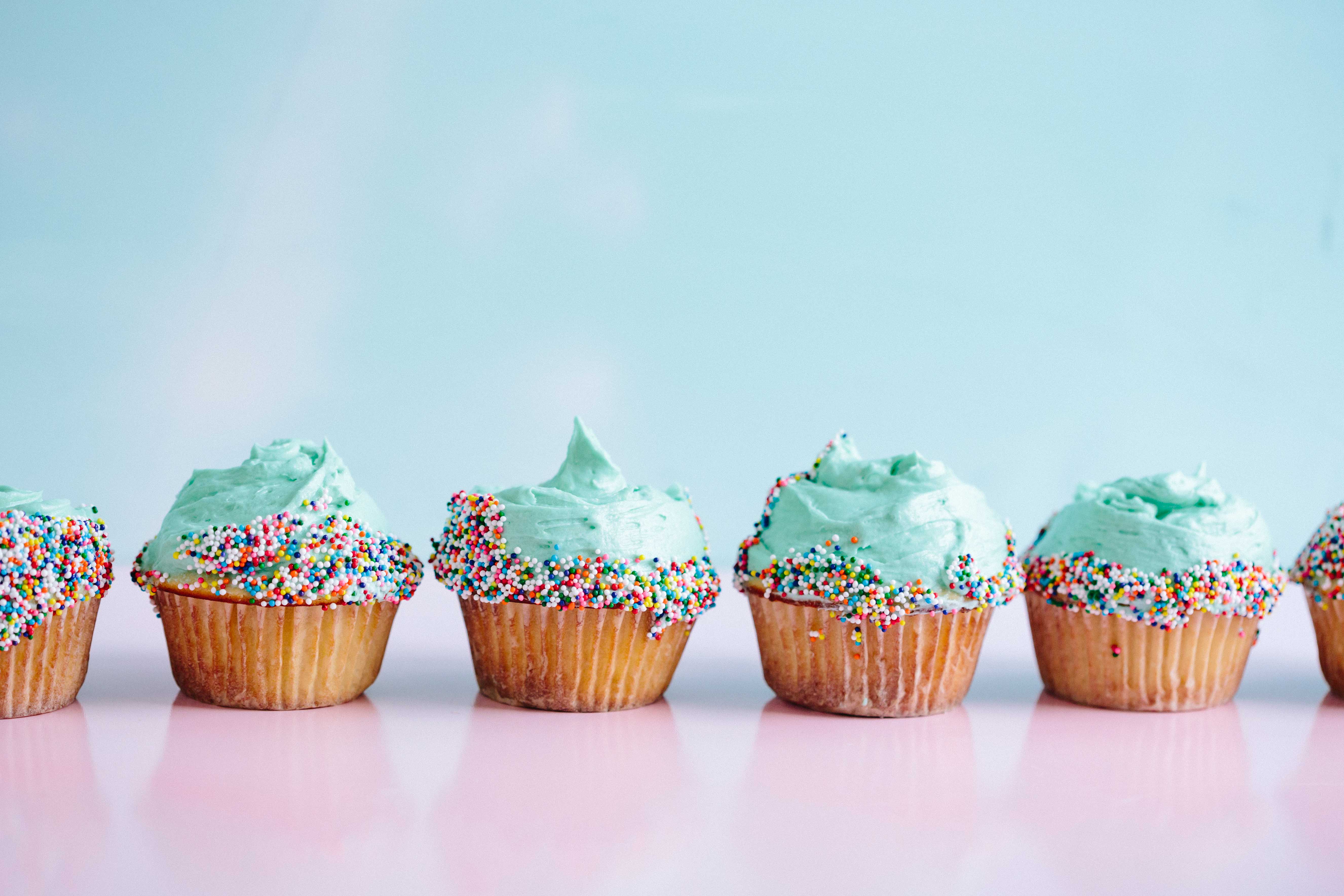 Six cupcakes with teal frosting and multicolored sprinkles on a pink surface.