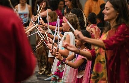 Children and adults in colorful traditional attire hold decorated sticks while dancing in a line indoors.