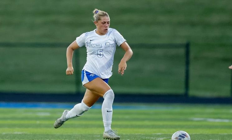 Female soccer player in white Thomas More jersey number 5 dribbling a ball on a grass field.