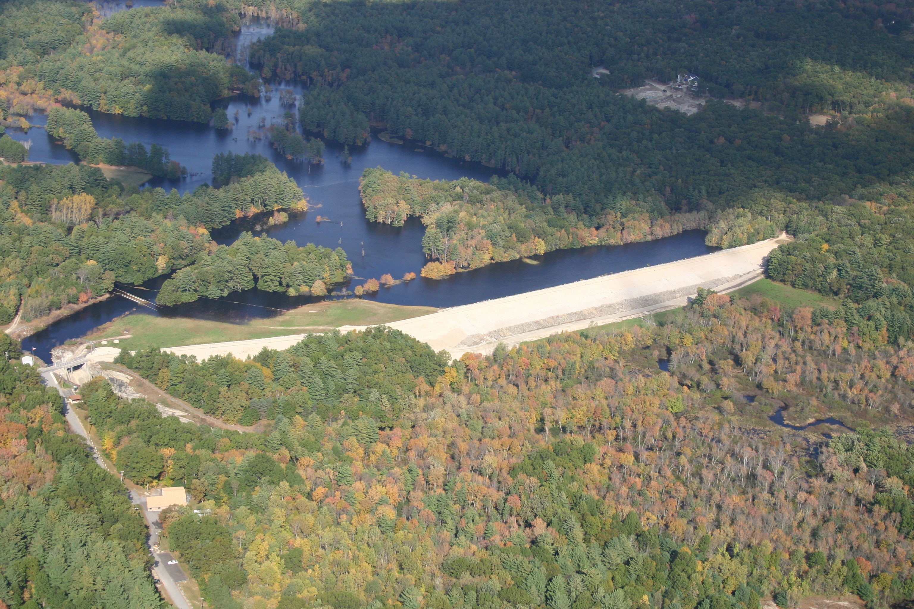 Aerial view of an earthen dam with a flooded reservoir surrounded by forested hills in early autumn.