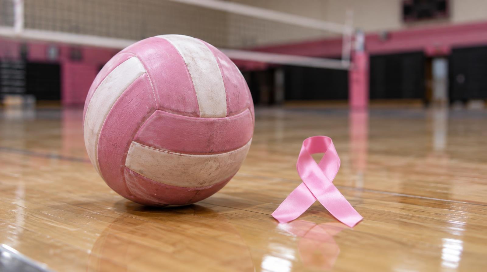 A pink volleyball rests on a hardwood gym floor beside a pink ribbon.