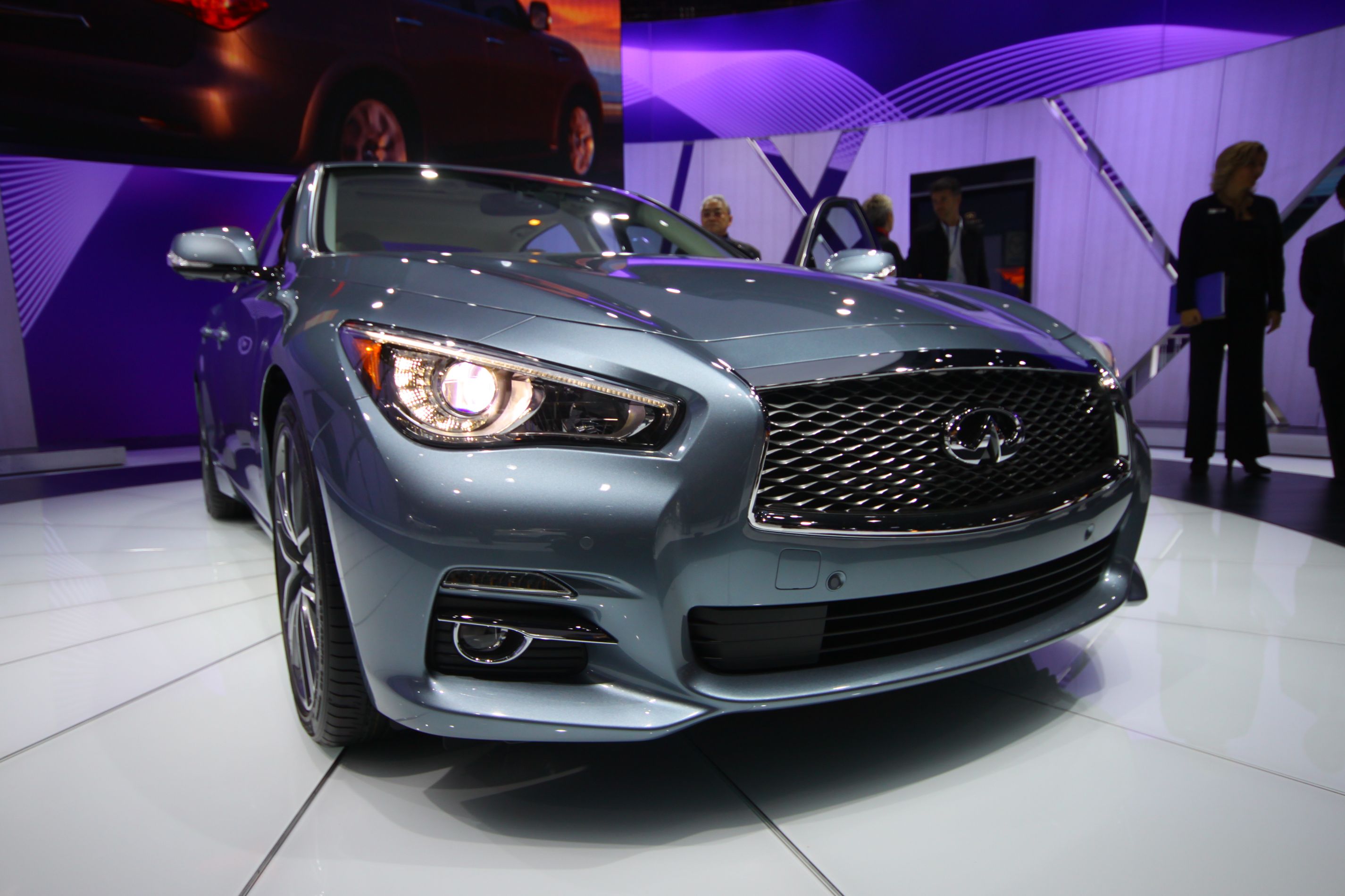Silver Infiniti Q50 sedan with headlights illuminated at an auto show.