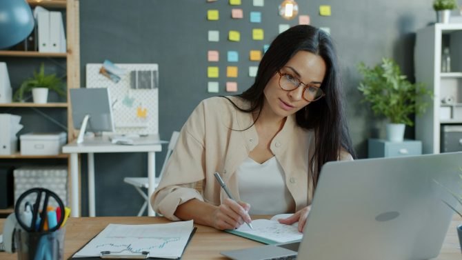 Woman wearing glasses writes in a notebook at a desk with an open laptop and colorful sticky notes on the wall behind her.