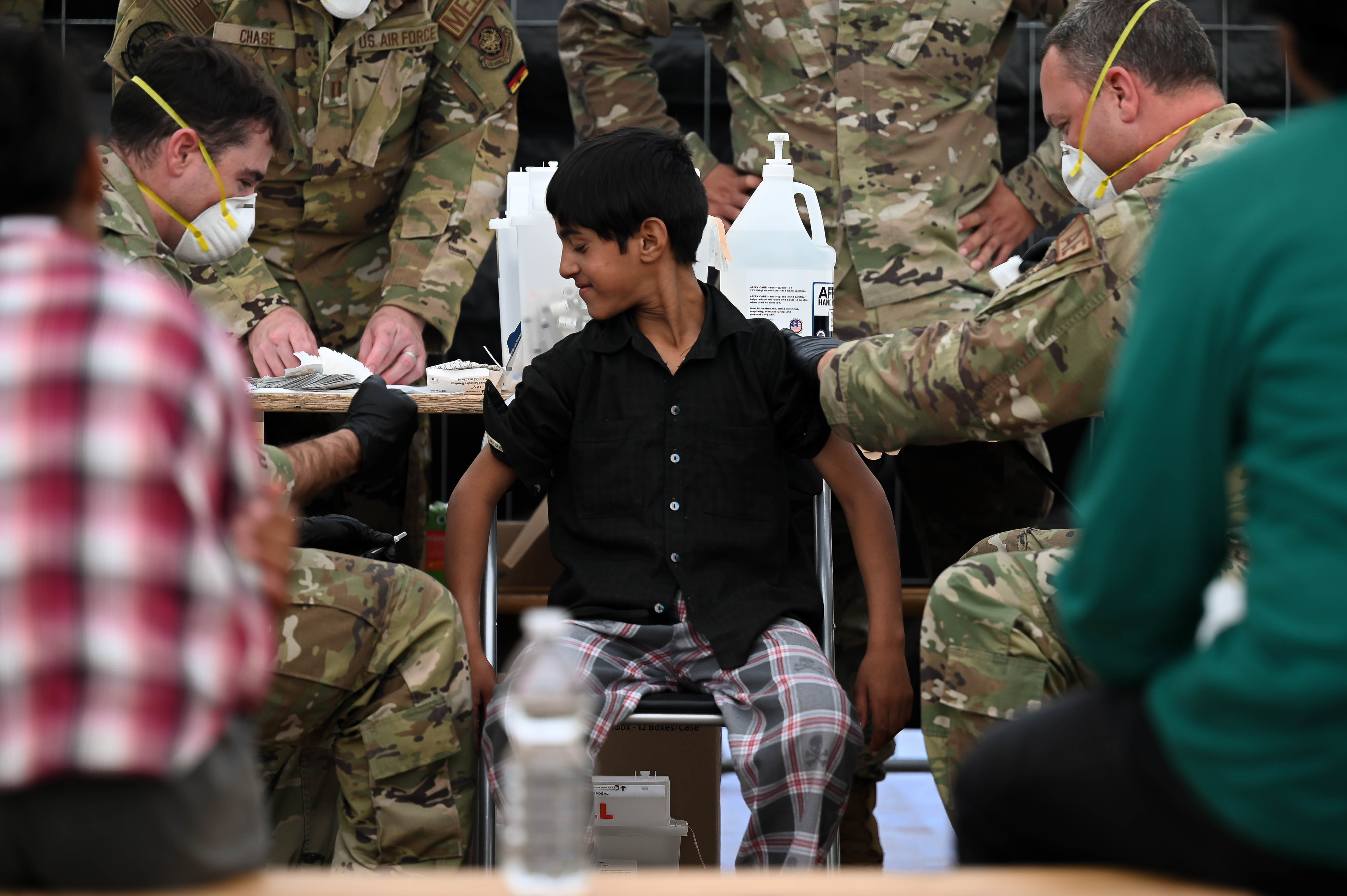 Two masked uniformed workers administer a vaccine to a calm young boy seated in a chair.