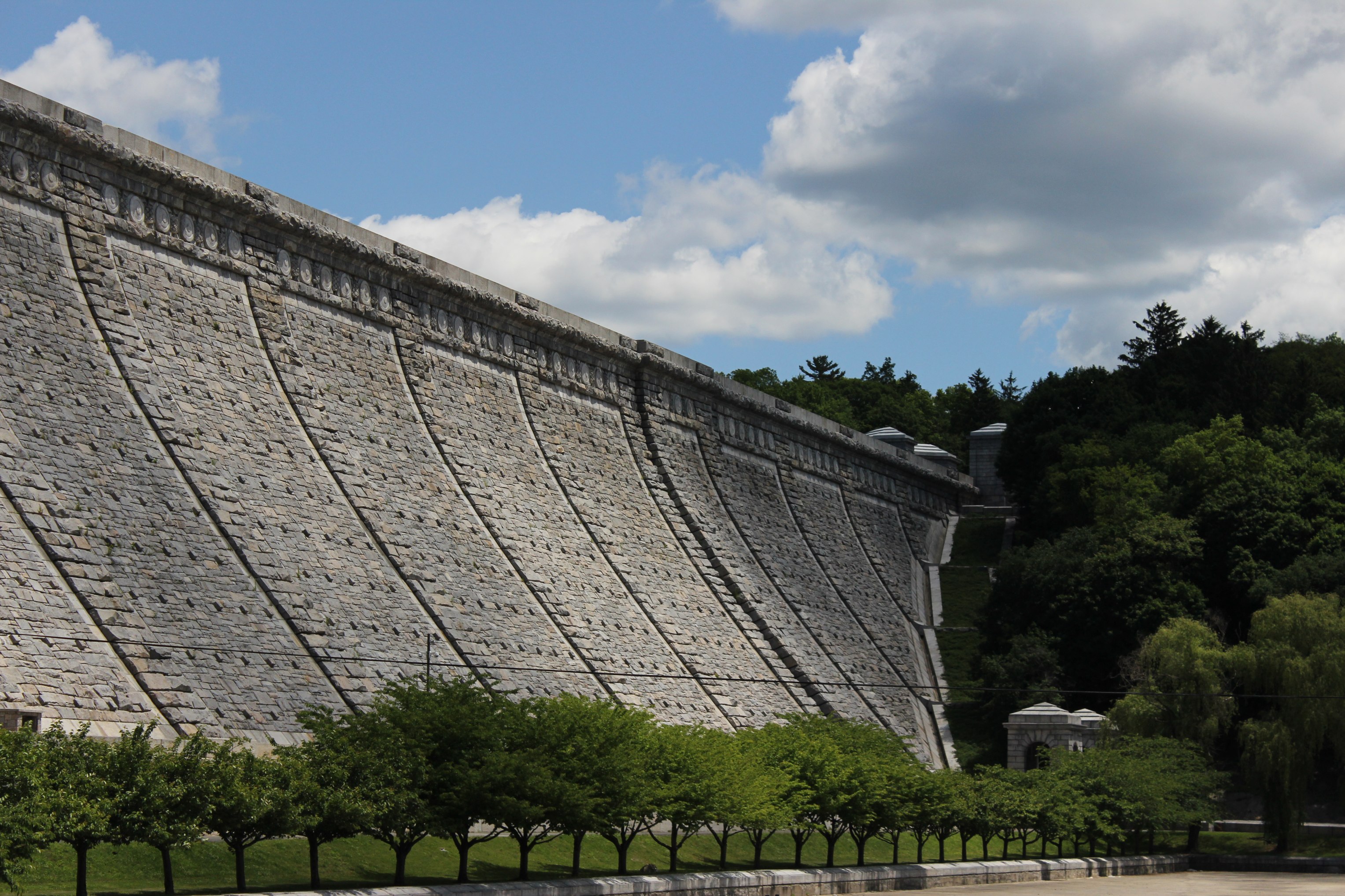 Large stone dam wall with diagonal buttress patterns viewed from below with trees at its base.