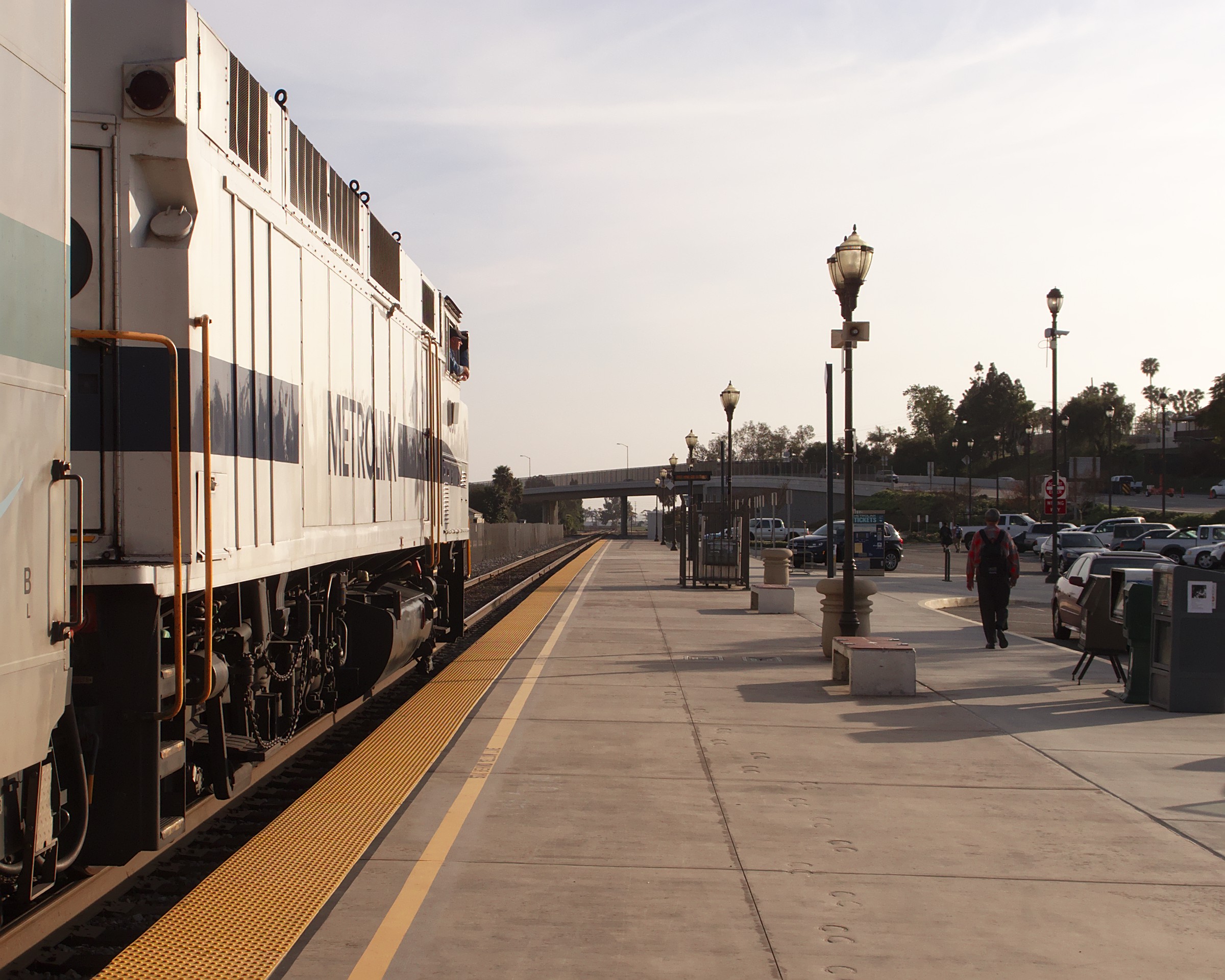 White Metrolink locomotive at a station platform with lamp posts and an overpass bridge.