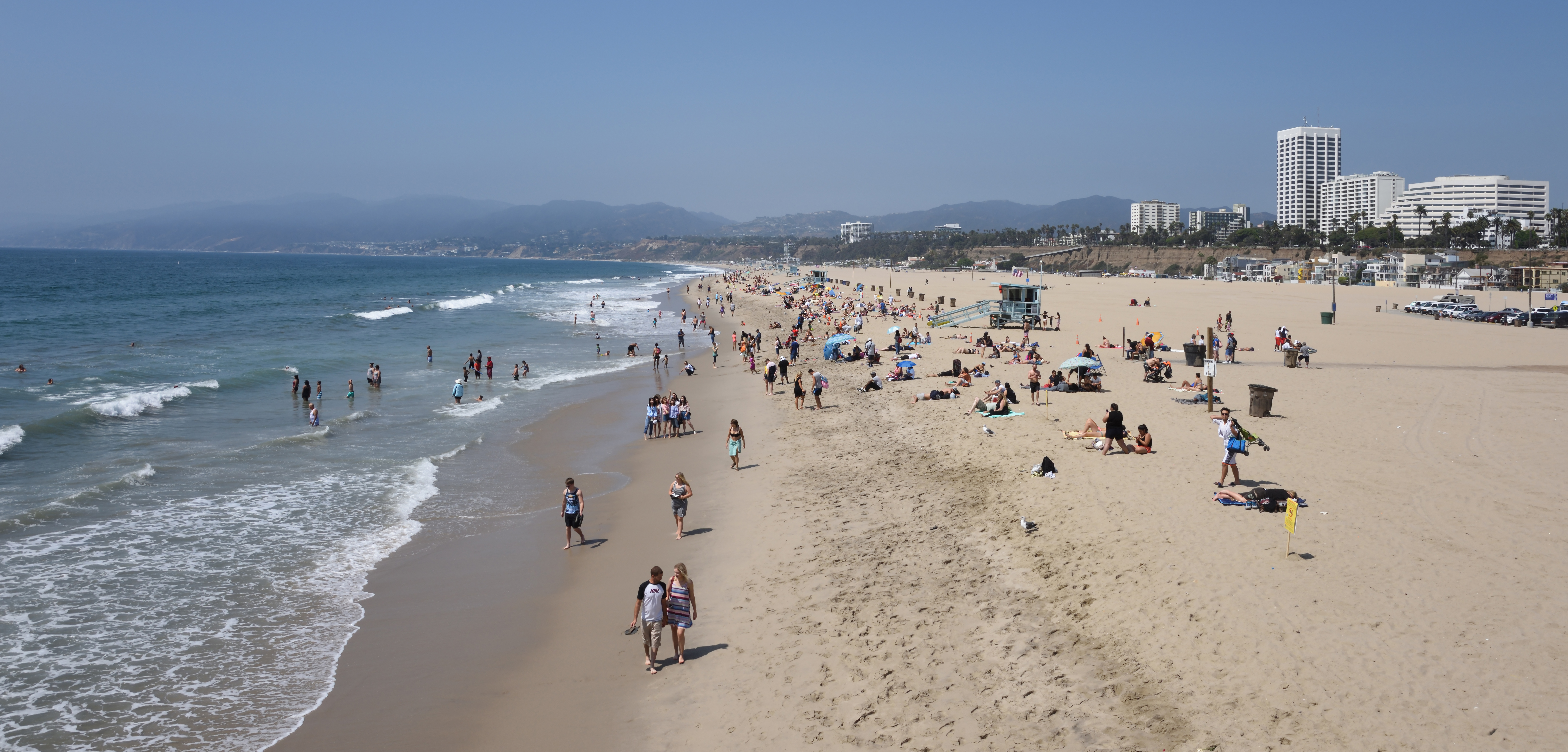 Crowded sandy beach with ocean waves, a lifeguard tower, and high-rise buildings in the background.