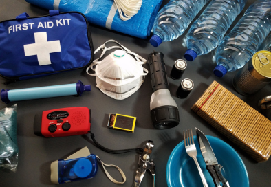 Flat-lay of emergency supplies including a first aid kit, water bottles, flashlight, batteries, mask, and radio.