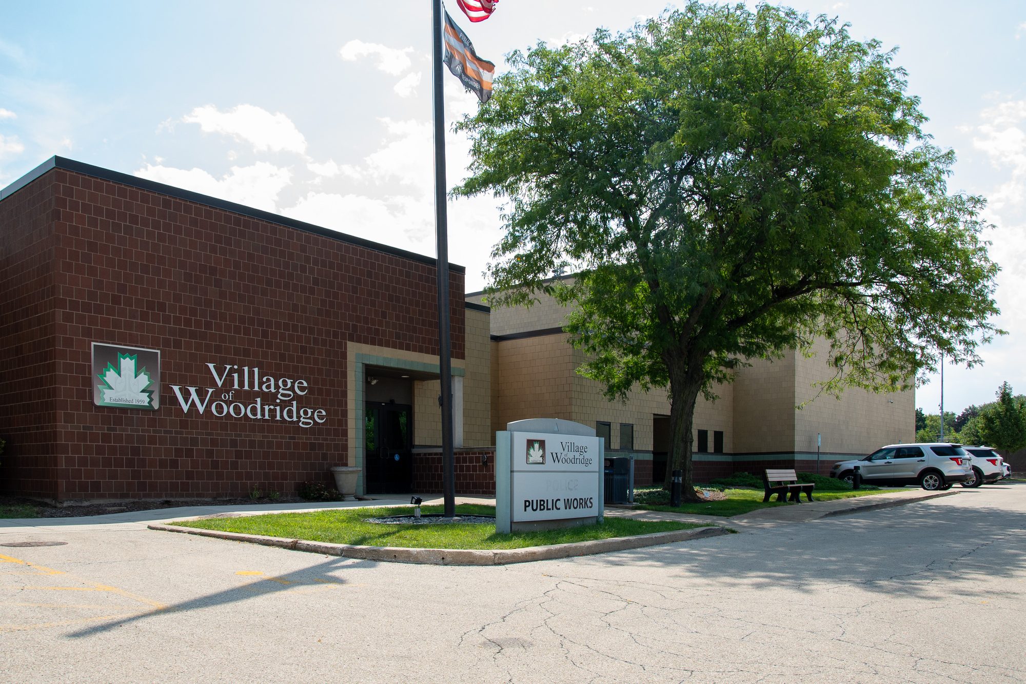 Brick municipal public works building with flagpole and large tree outside.