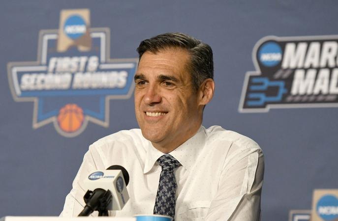 Man in white dress shirt and dark tie smiling at a press conference with NCAA March Madness signage behind him.