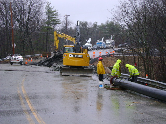 Workers in high-visibility gear and an excavator handle a large pipe on a wet, closed road.