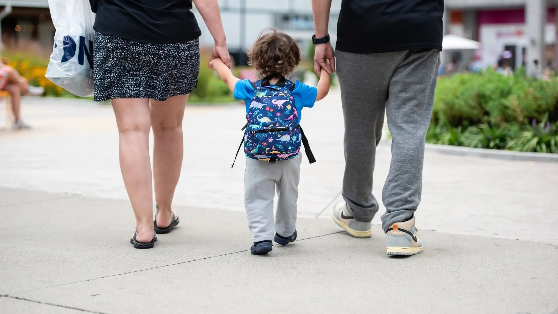 Toddler in a blue dinosaur backpack walks between two adults on an outdoor path.
