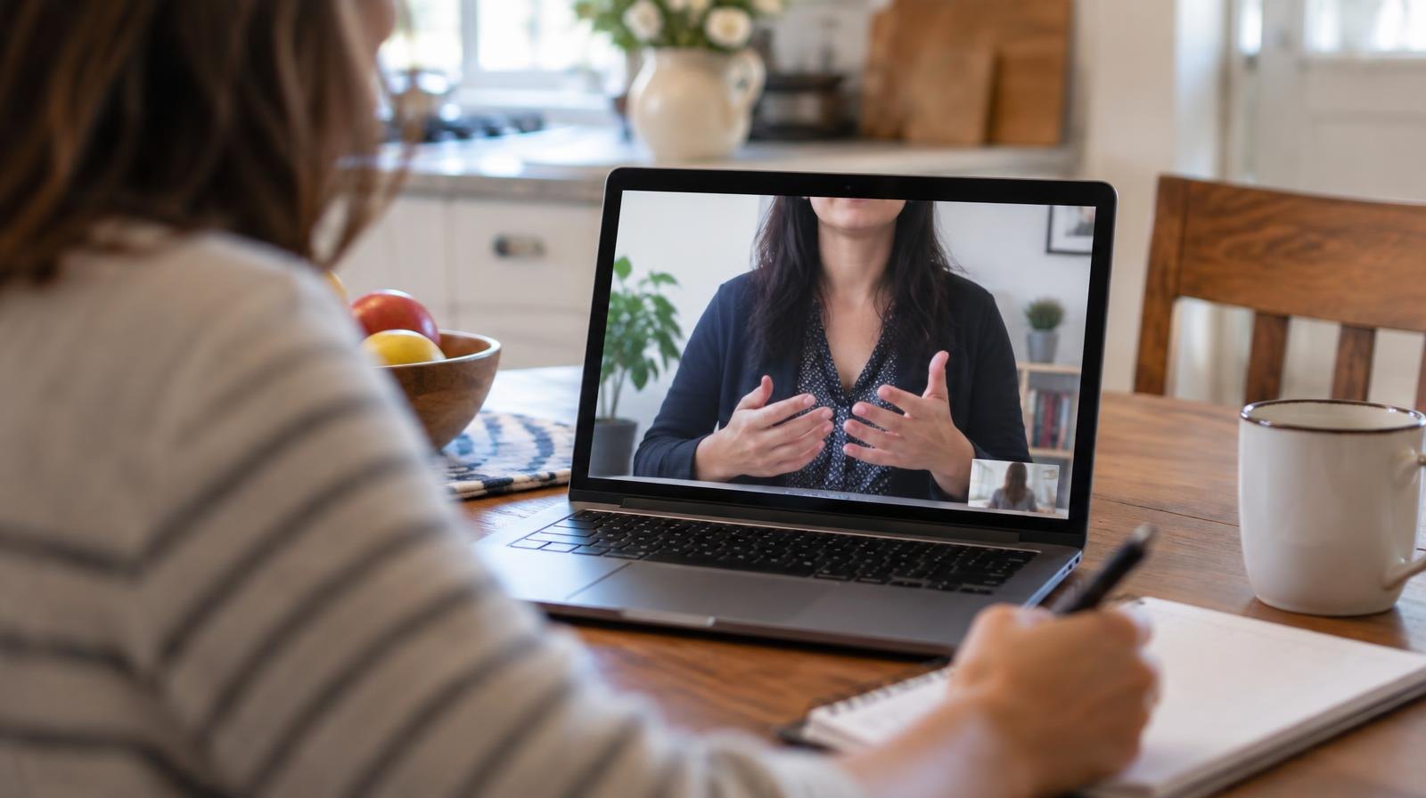 Open laptop on a table showing a video call, with a notebook and pen beside it.