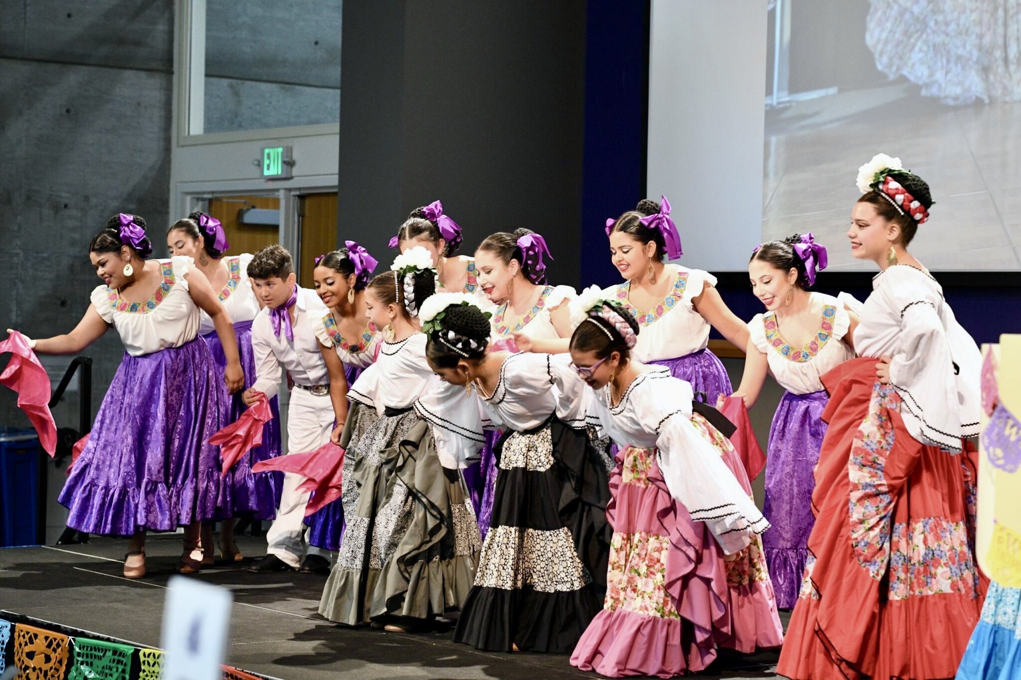 Young women and girls in colorful tiered skirts perform a folkloric dance on a stage.
