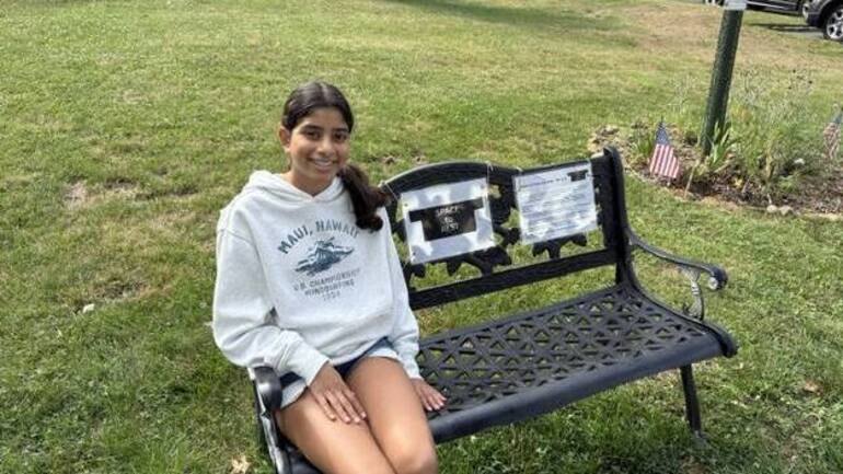 A teenage girl in a gray sweatshirt sits on the grass next to a black metal bench with signs attached.