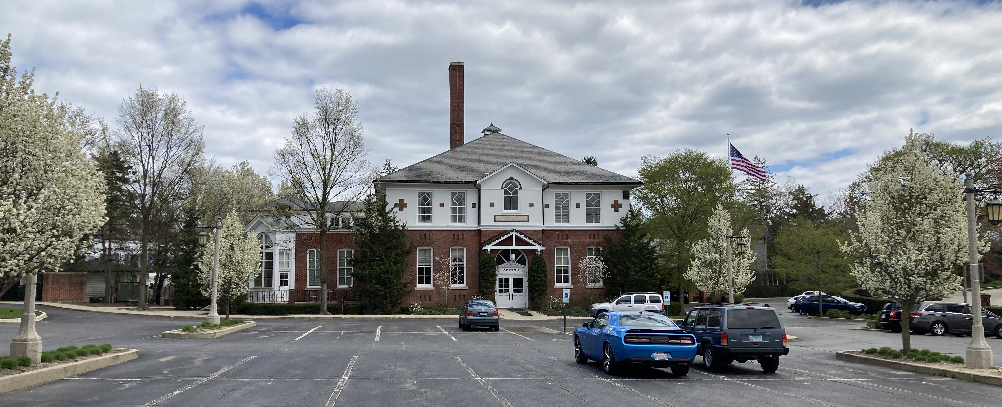 Two-story brick building with American flag and white-flowering trees in front.