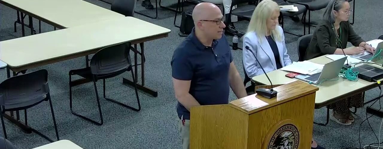 A man speaks at a podium in a municipal meeting room while two women sit at a table behind him.