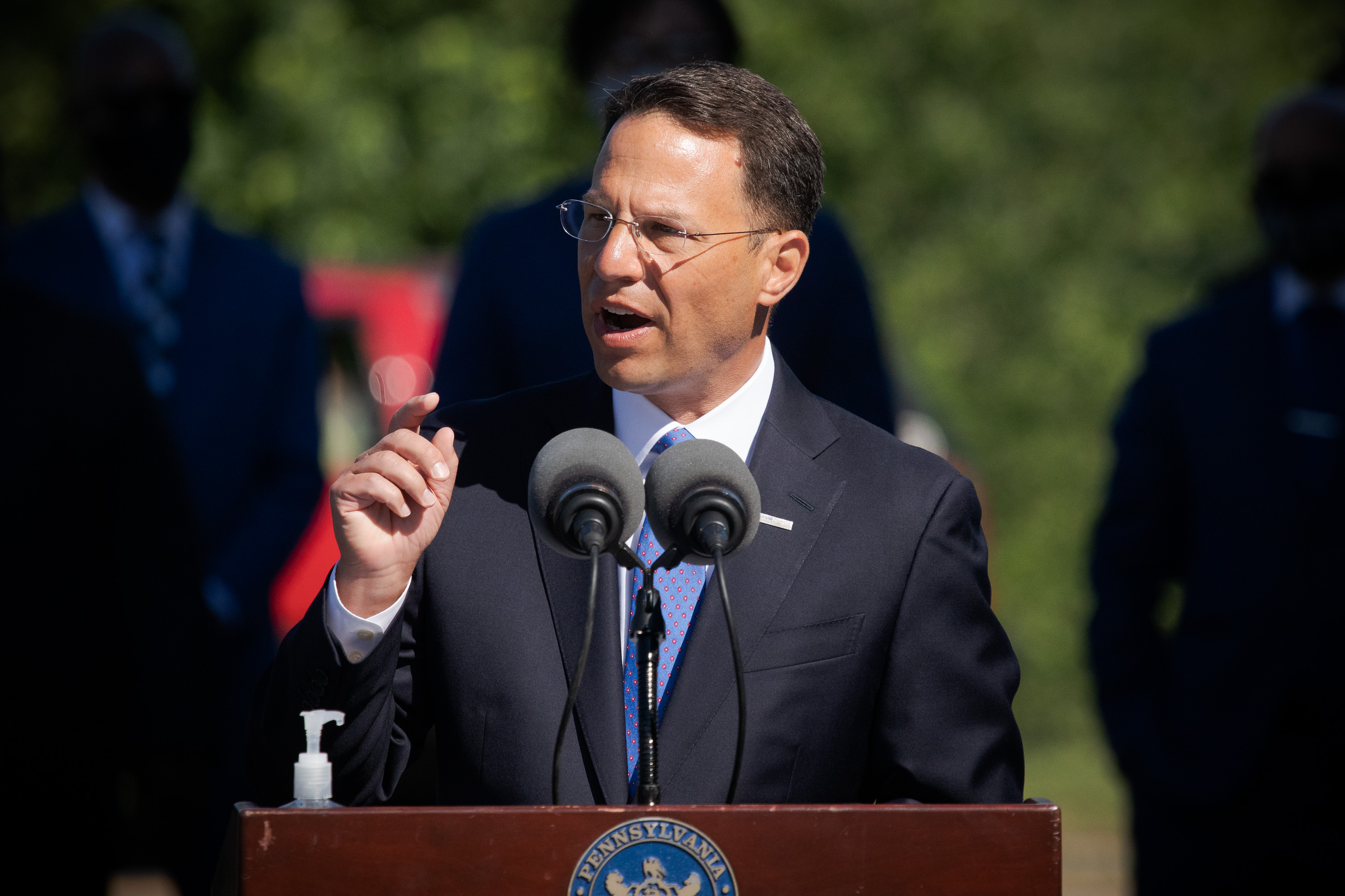 Man in dark suit and blue tie at an outdoor podium bearing the Pennsylvania state seal.
