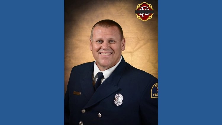 Man in dark navy fire department dress uniform with badge, smiling at camera.