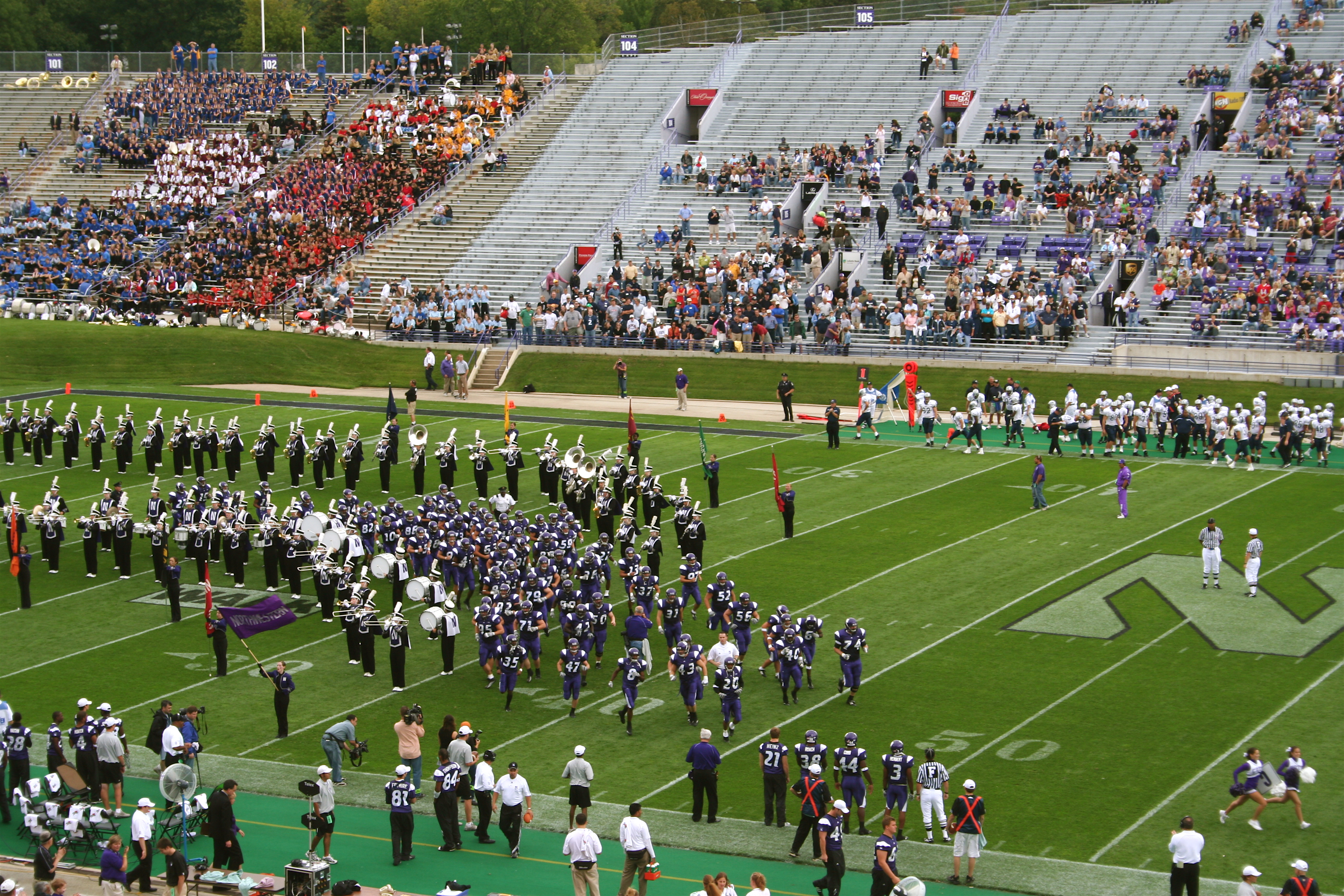 Northwestern football players in purple uniforms and marching band on a stadium field with fans in the stands.