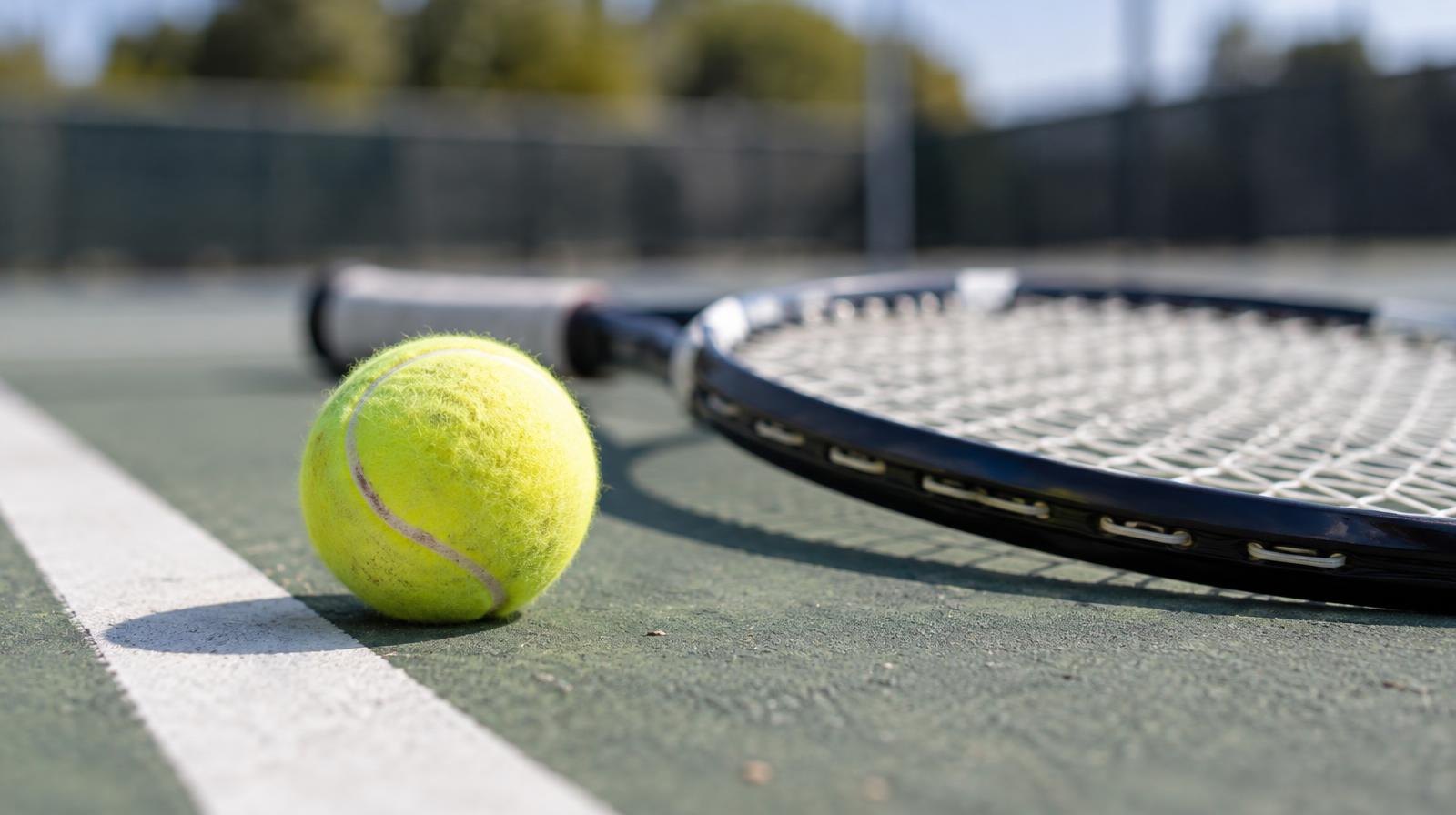 A yellow tennis ball rests on a green hard court beside a white baseline and a tennis racket.