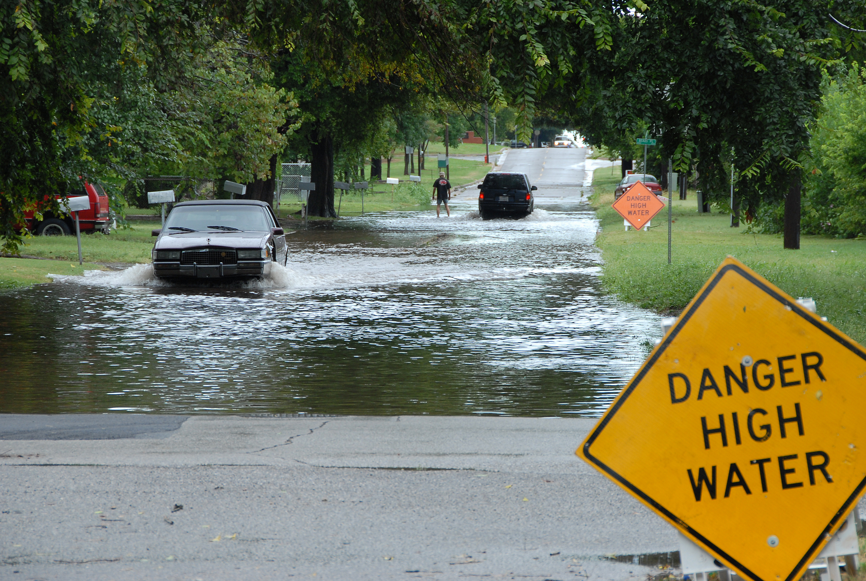 Cars drive through a flooded street past a yellow Danger High Water warning sign.